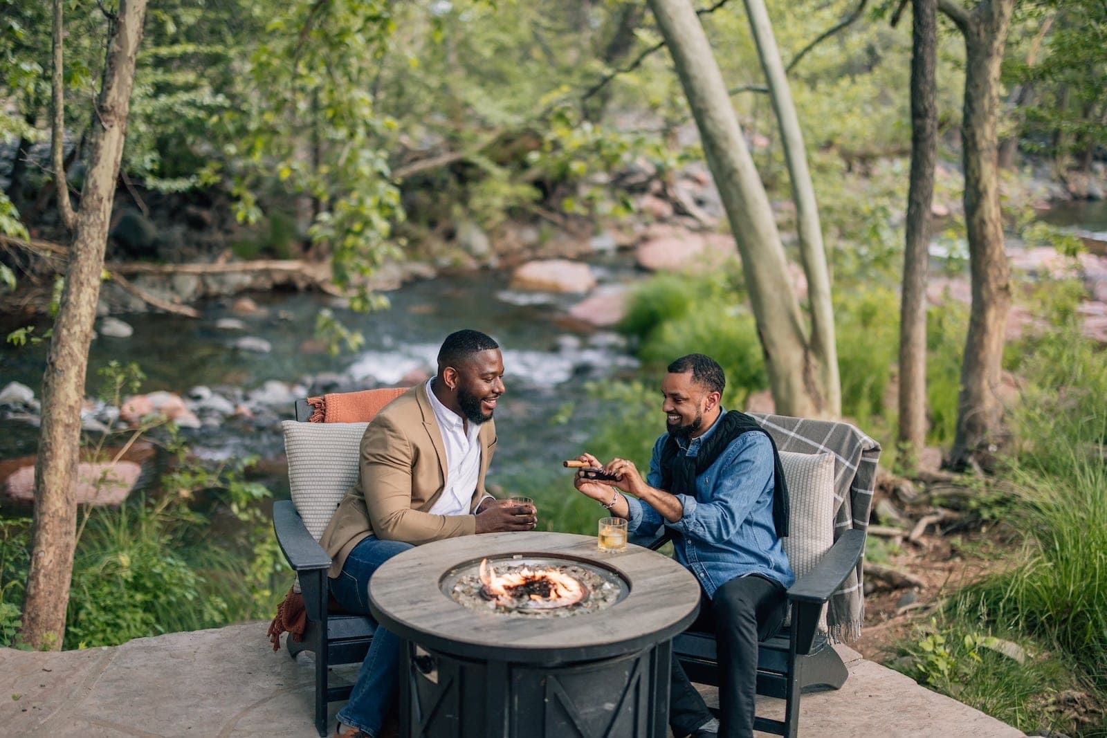 Two men sitting by a fire pit next to a creek.