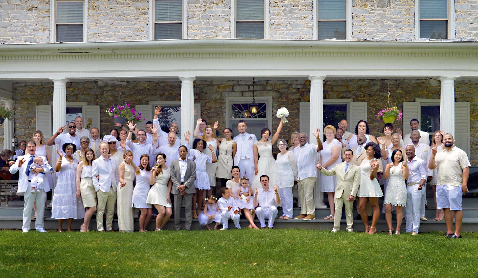 Wedding guests in white attire pose on the lawn in front of a stone building with a porch.