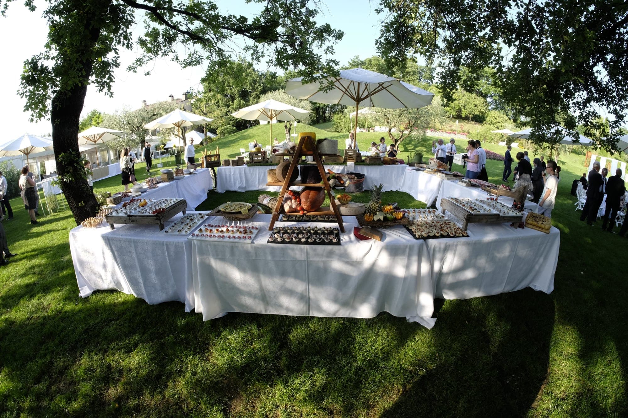 Buffet tables are set up on a grassy lawn under trees with white umbrellas.