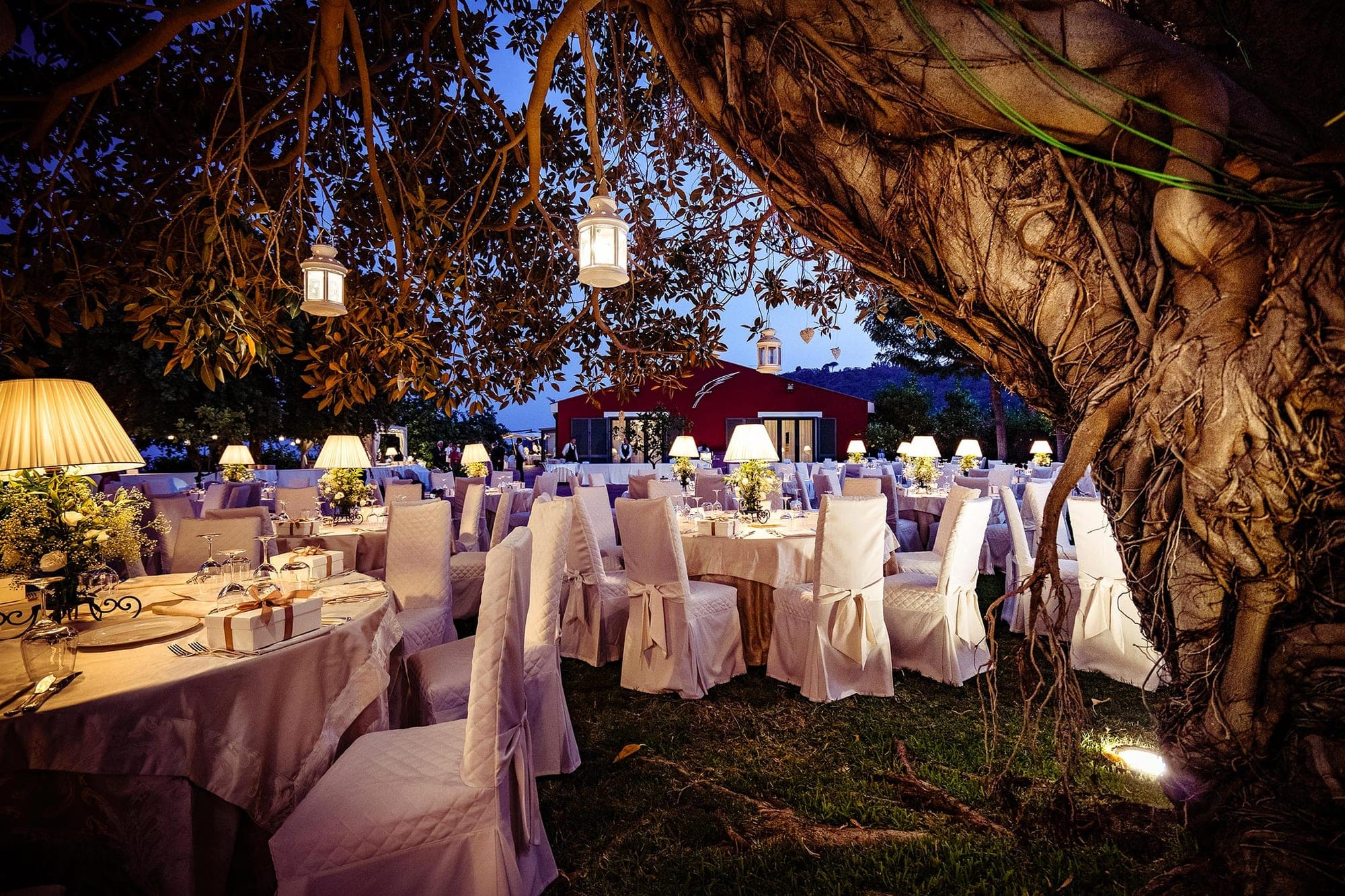 Outdoor reception tables set for a wedding under a large tree at dusk.