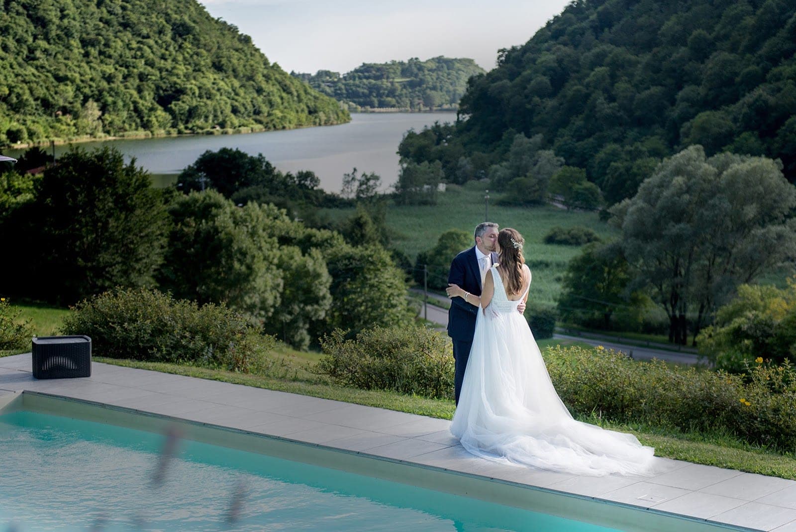 A couple kisses by a pool with a lake and hills in the background.