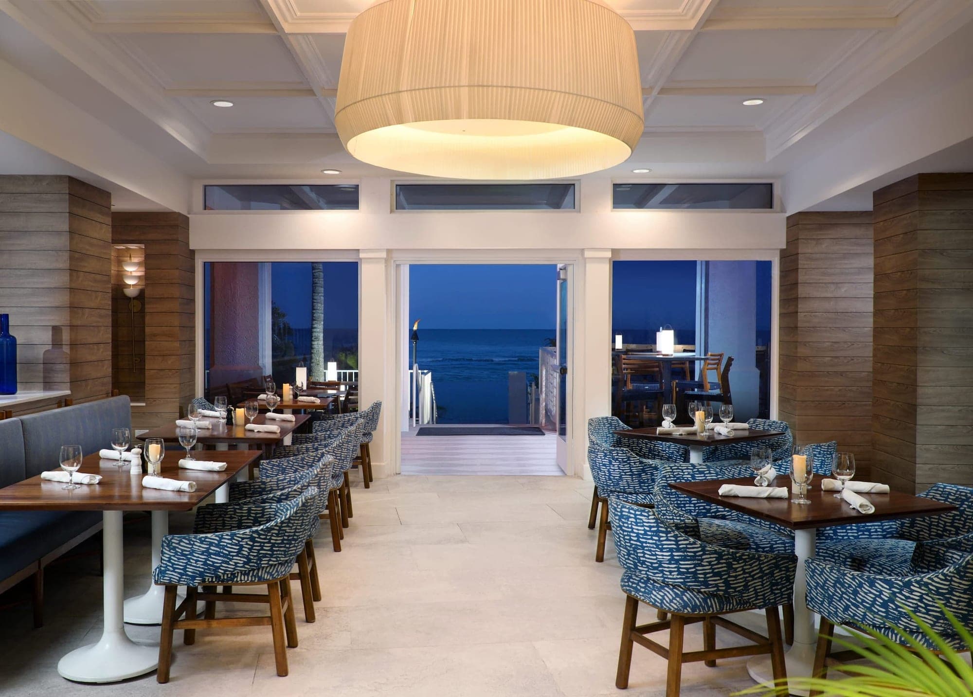Restaurant dining room with tables set for dinner and a view of the ocean at dusk.