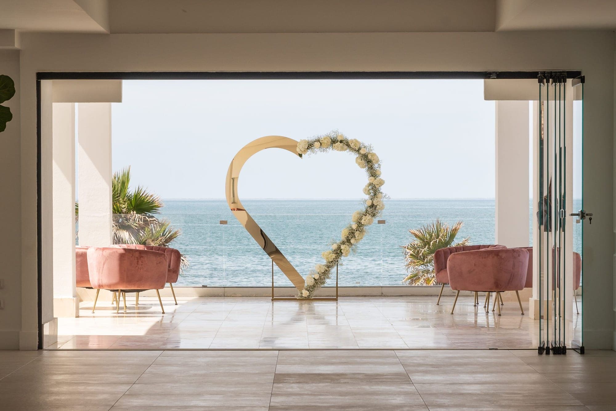 A heart-shaped floral arch stands before a sea view, with two pink chairs on either side.