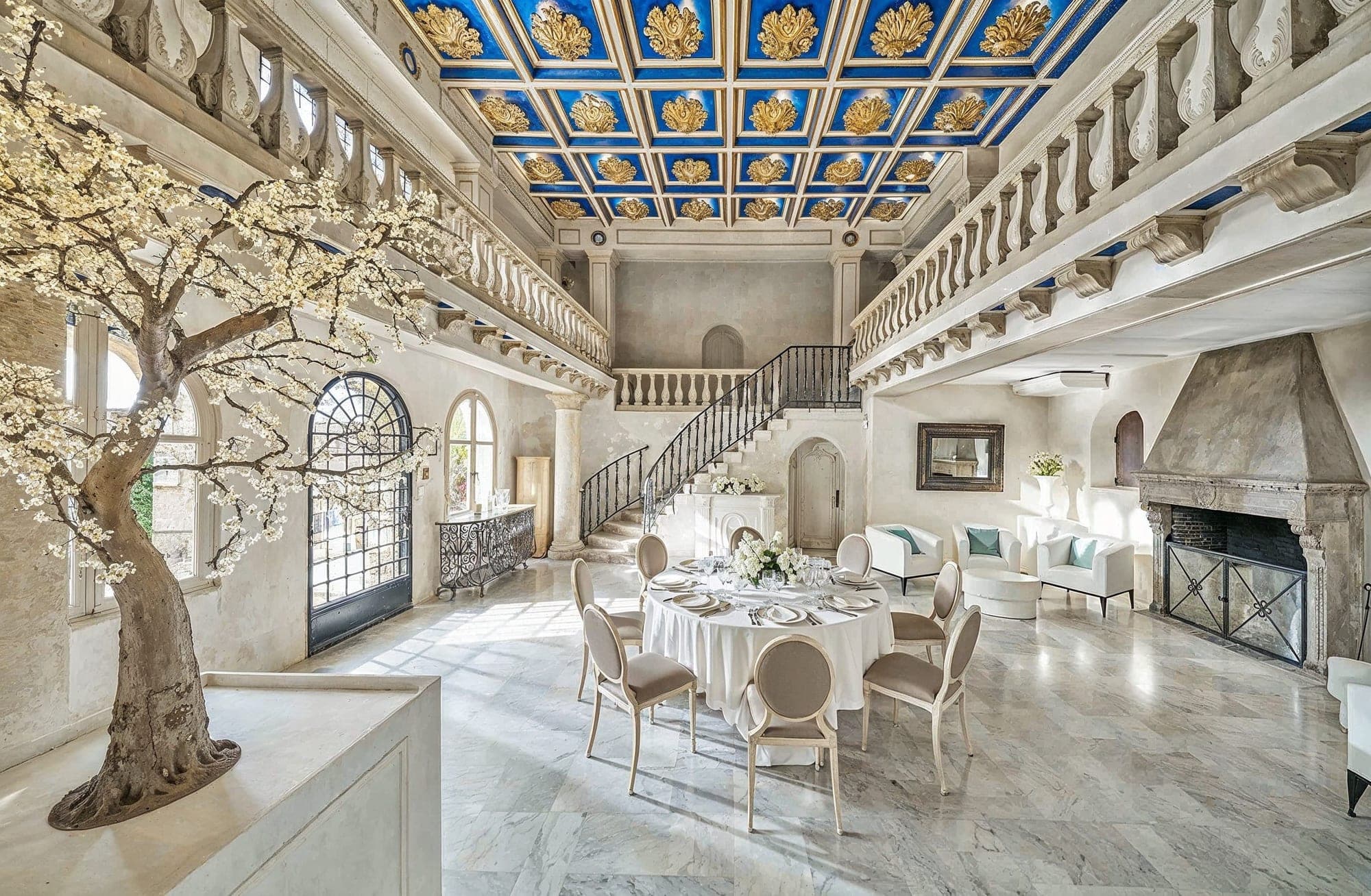A round dining table is set for a meal in a large hall with a grand staircase and ornate ceiling.