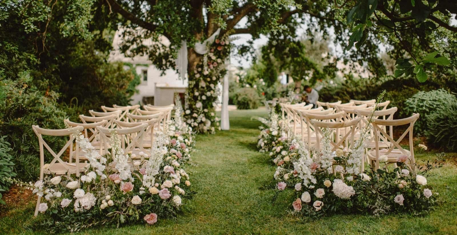 Outdoor wedding ceremony setup with chairs and flowers under a large tree.