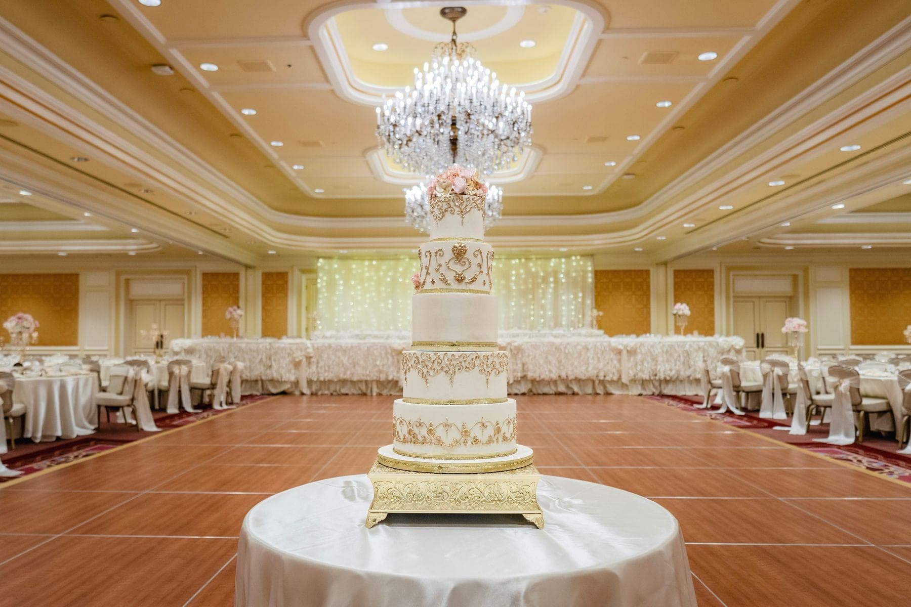 A tall wedding cake sits on a table in a reception hall with tables and chairs set up.