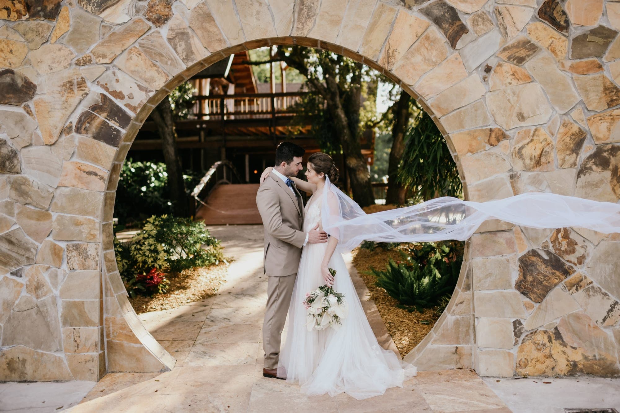A bride and groom stand embracing under a stone archway, with their wedding veil flowing in the wind.