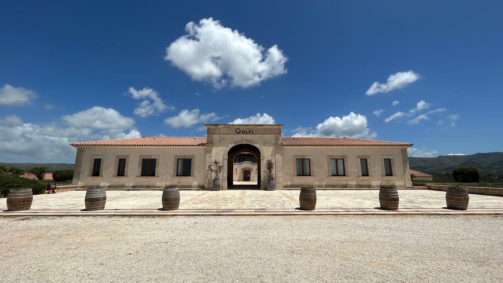 Exterior of a long, light-colored building with an arched entrance and barrels in the foreground.
