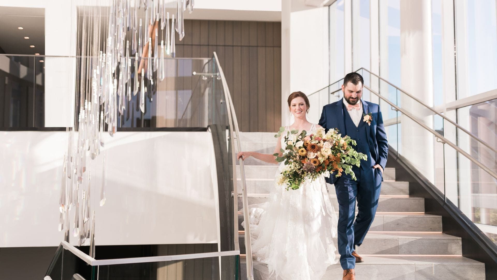 A bride and groom walk down a modern staircase with a chandelier.