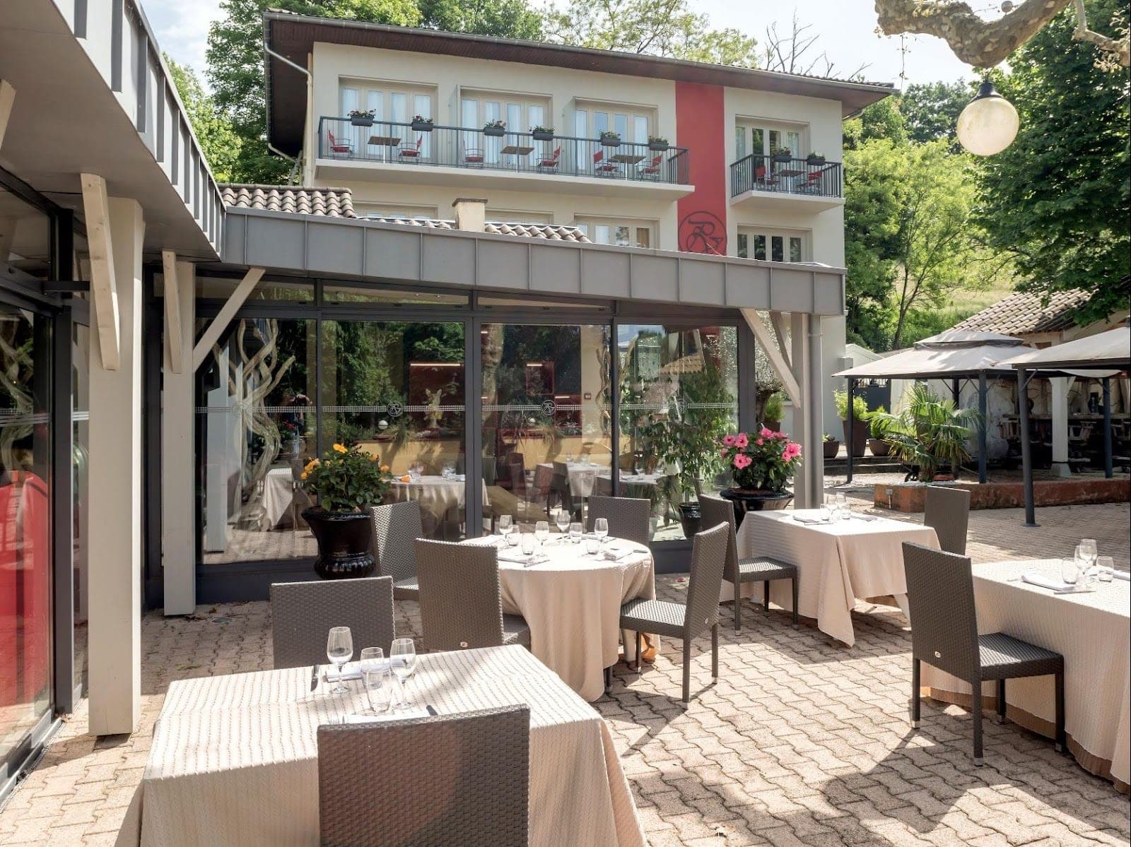 Tables set for dining on a paved terrace outside a building with balconies.