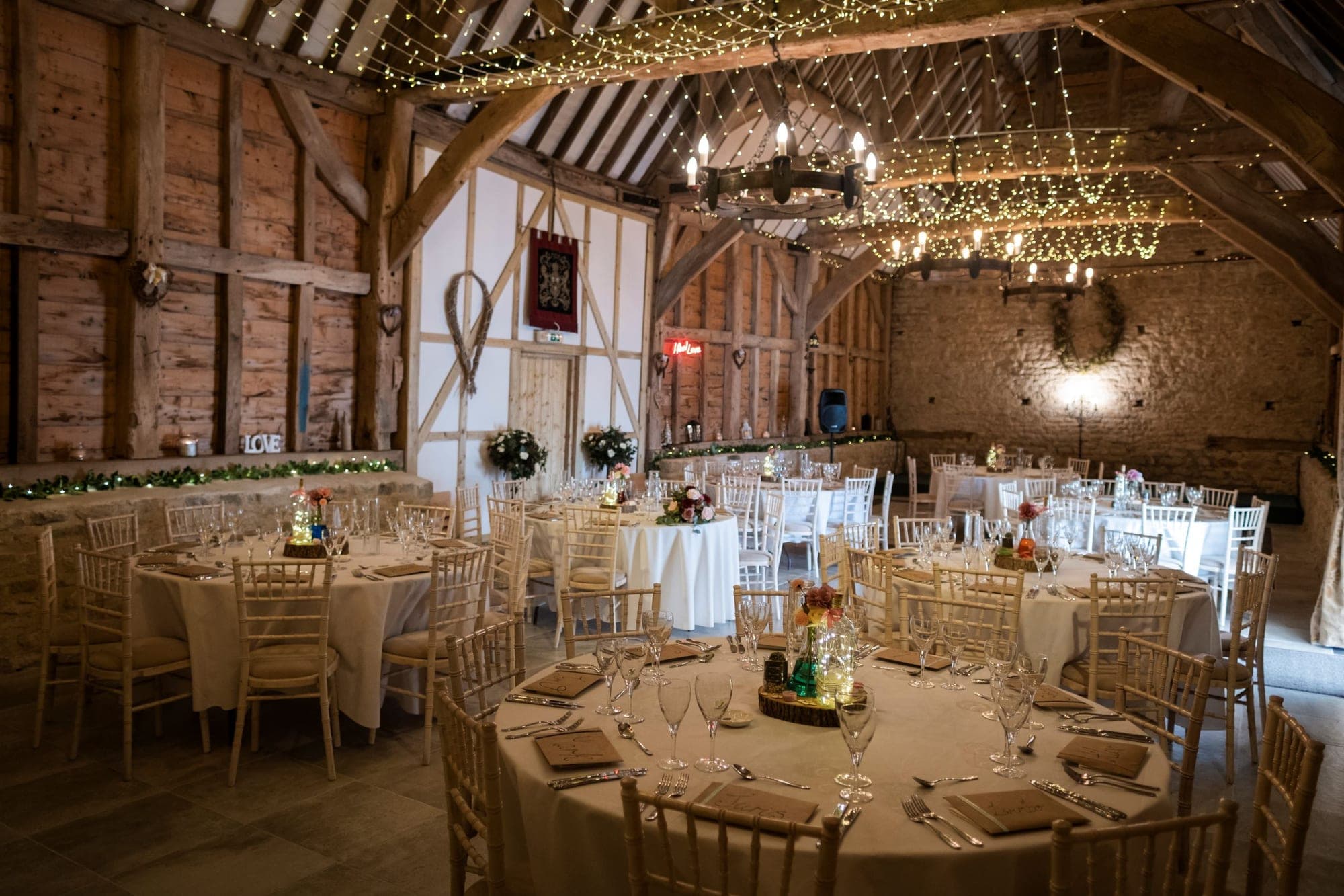 A wedding reception set up in a barn with round tables and chairs, decorated with fairy lights.