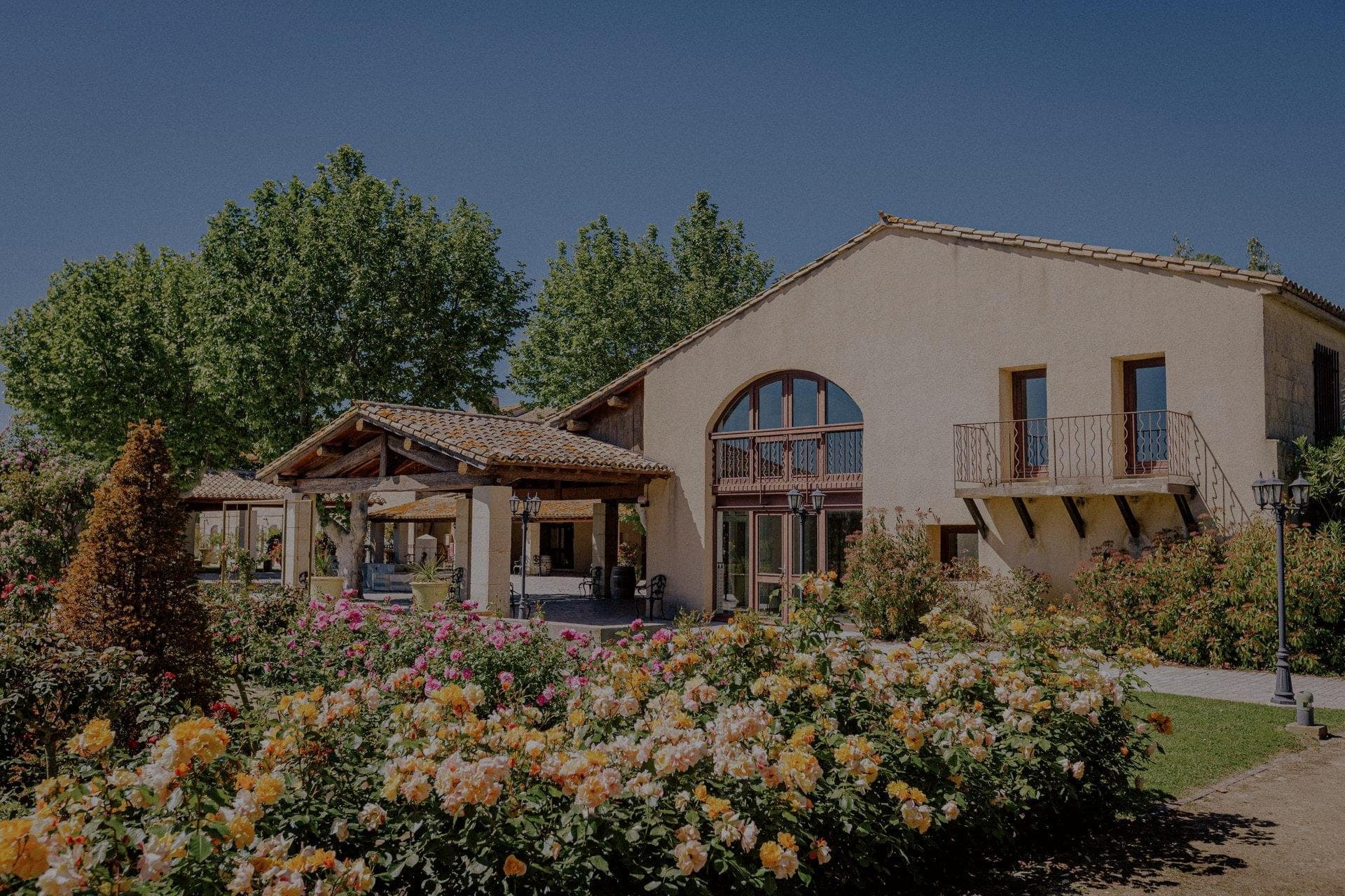 Exterior of a building with a large arched window and a balcony, surrounded by blooming roses and trees.