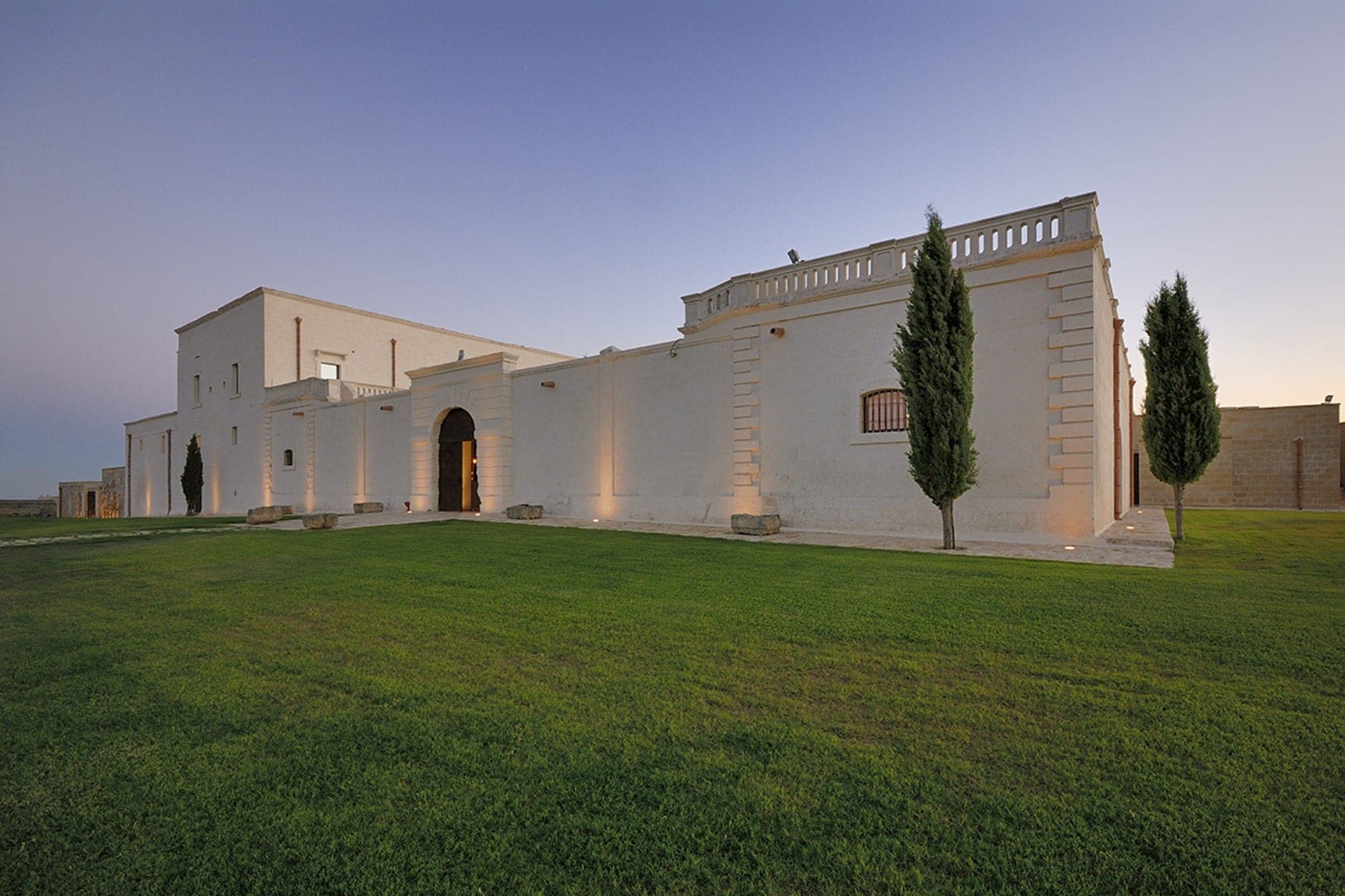 Exterior of a white building with an arched entrance and cypress trees in front of a lawn.
