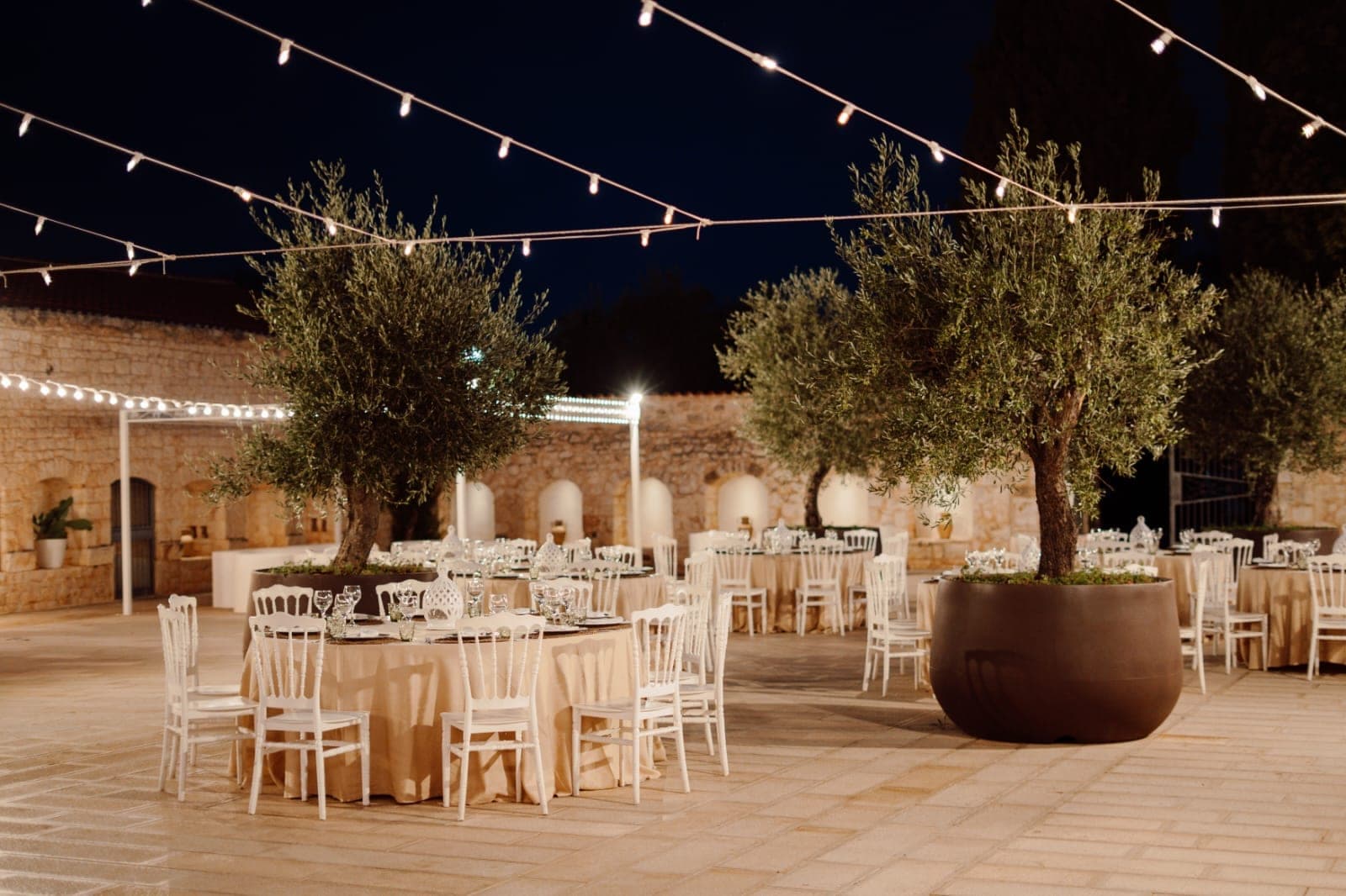 Outdoor reception area with tables, chairs, and olive trees under string lights at night.