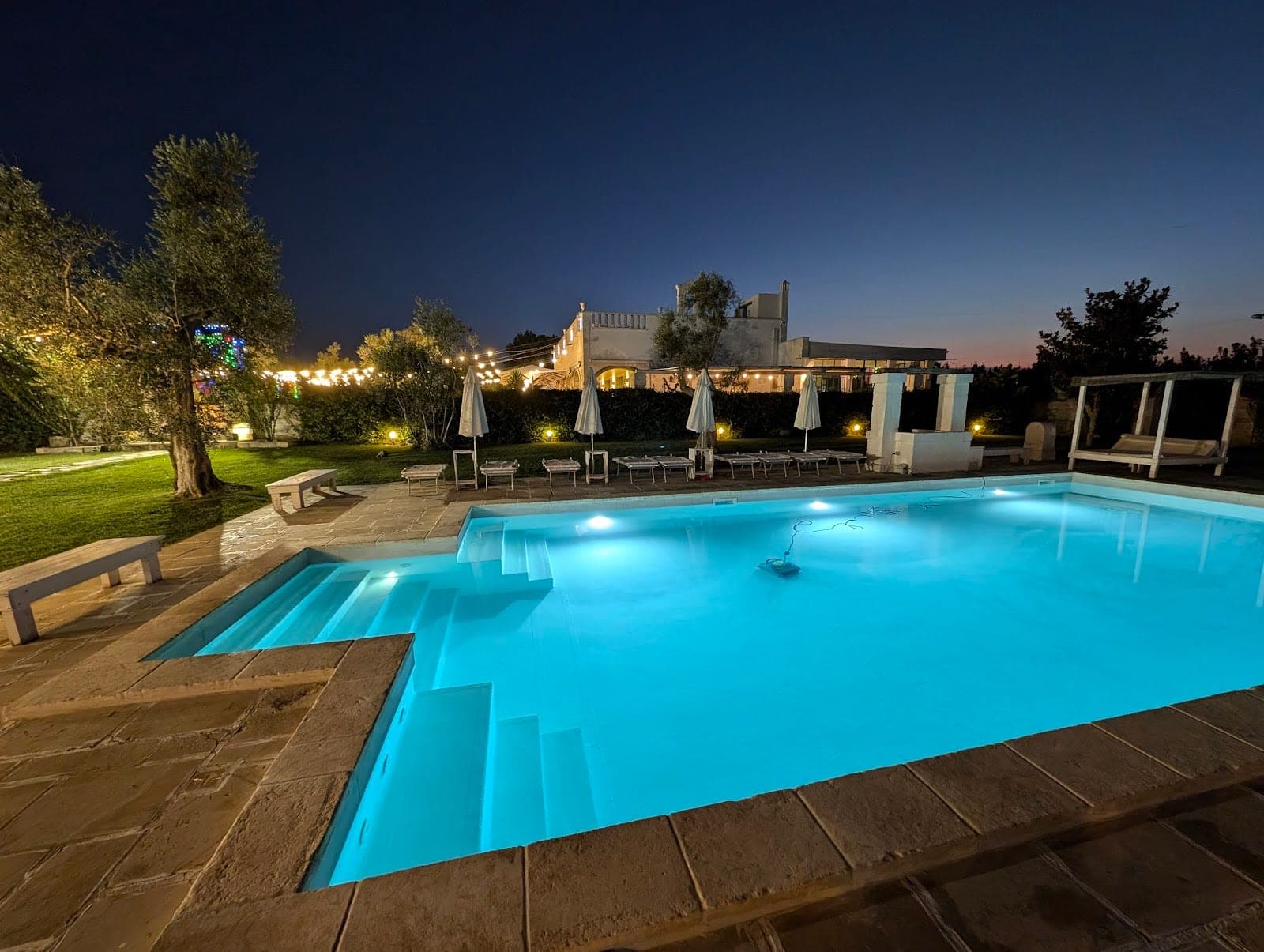 Swimming pool and venue exterior at dusk with lights and lounge chairs.