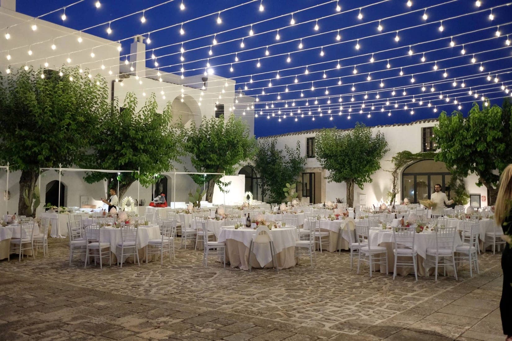Courtyard set for a wedding reception with tables, chairs, and string lights overhead.
