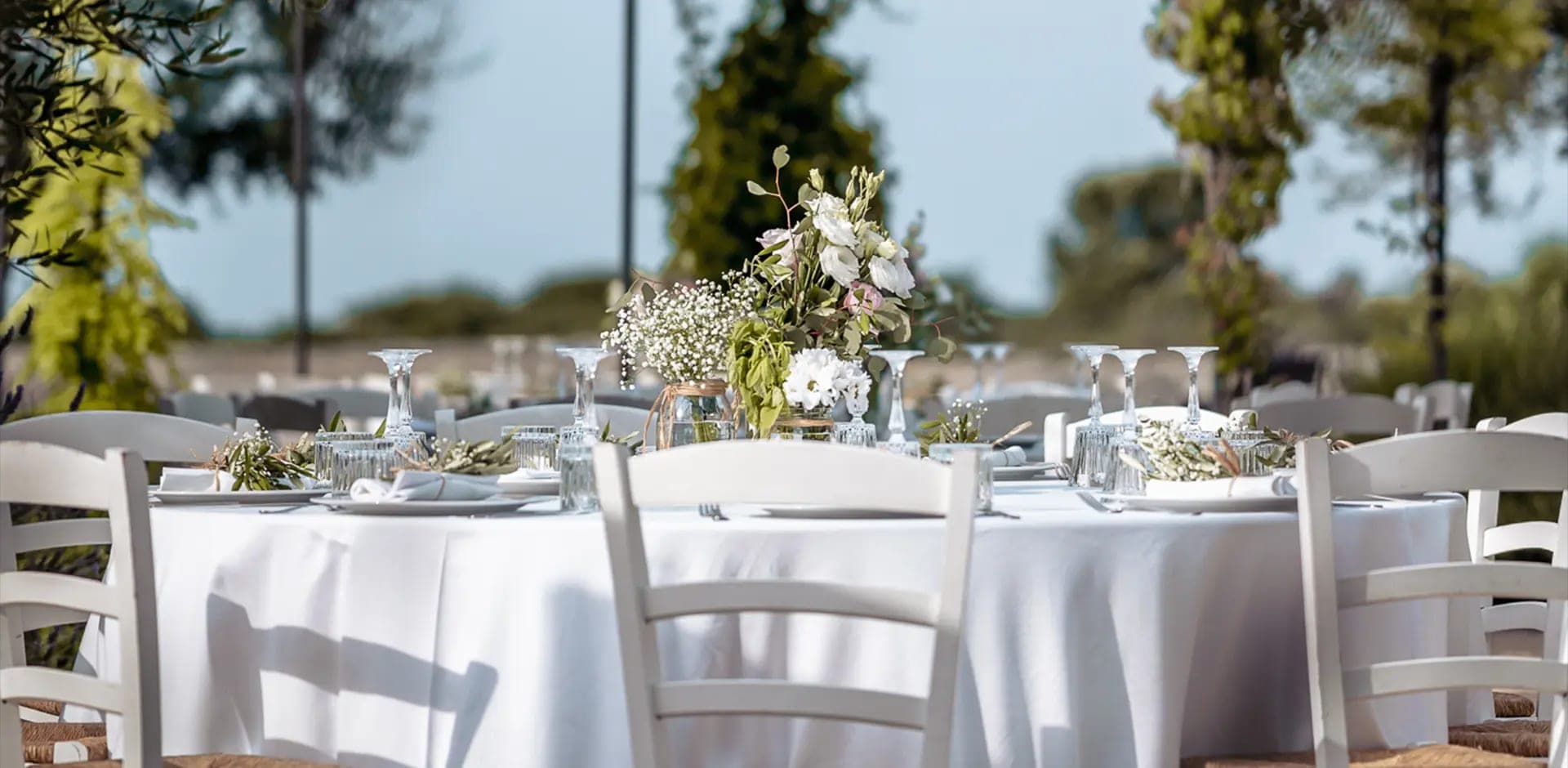 Outdoor wedding reception table setting with white chairs and floral centerpieces.