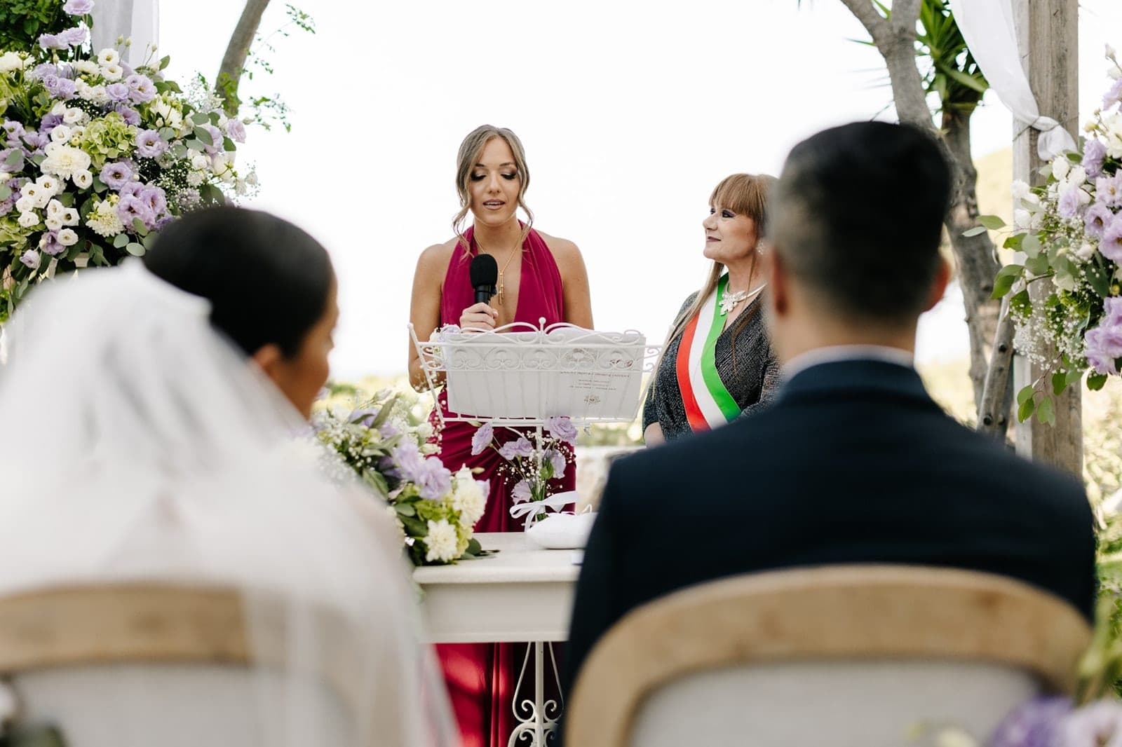 A wedding ceremony is taking place outdoors with floral decorations and officiants.