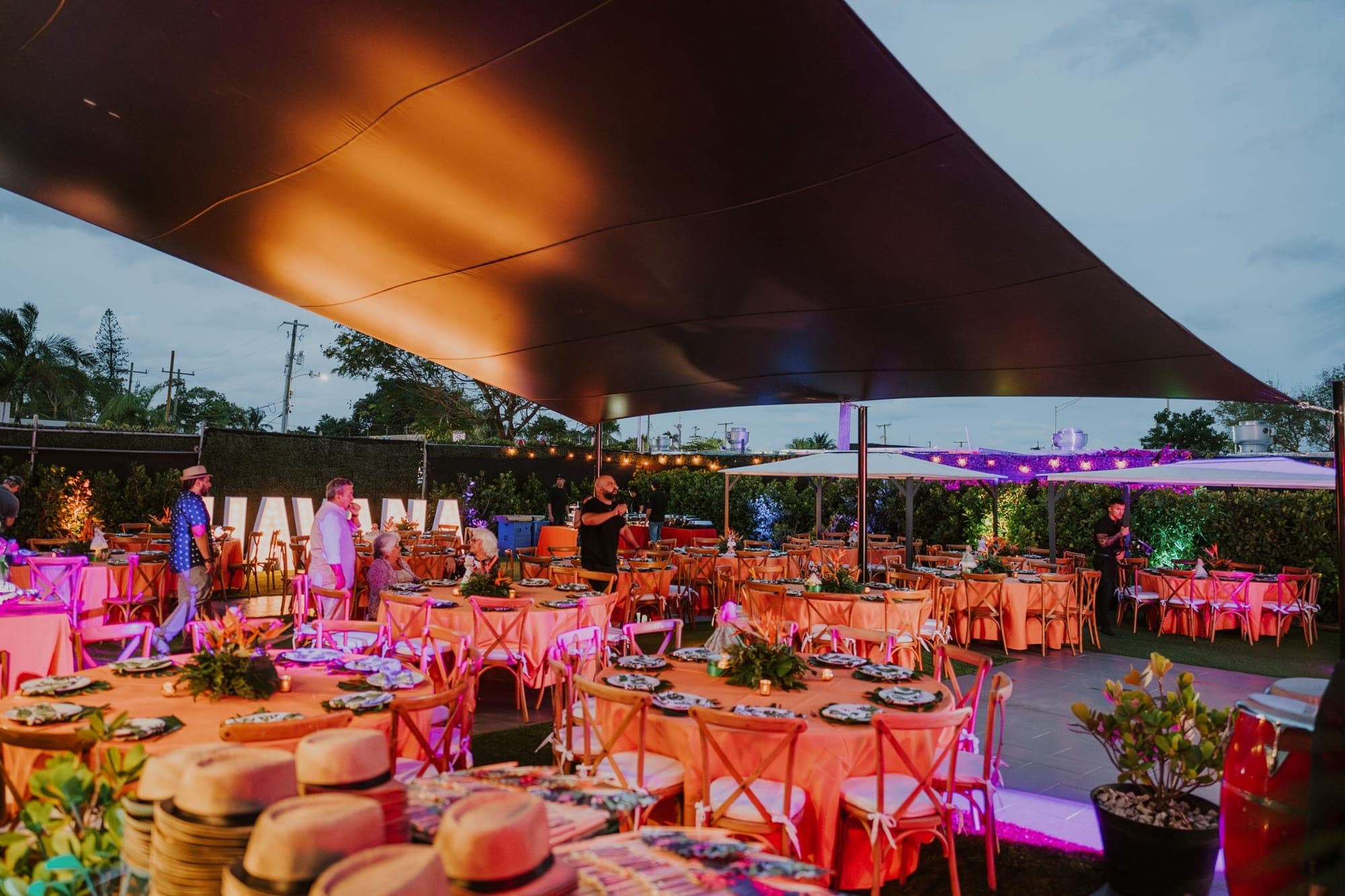 Outdoor reception tables set with orange tablecloths and chairs under a large dark canopy.