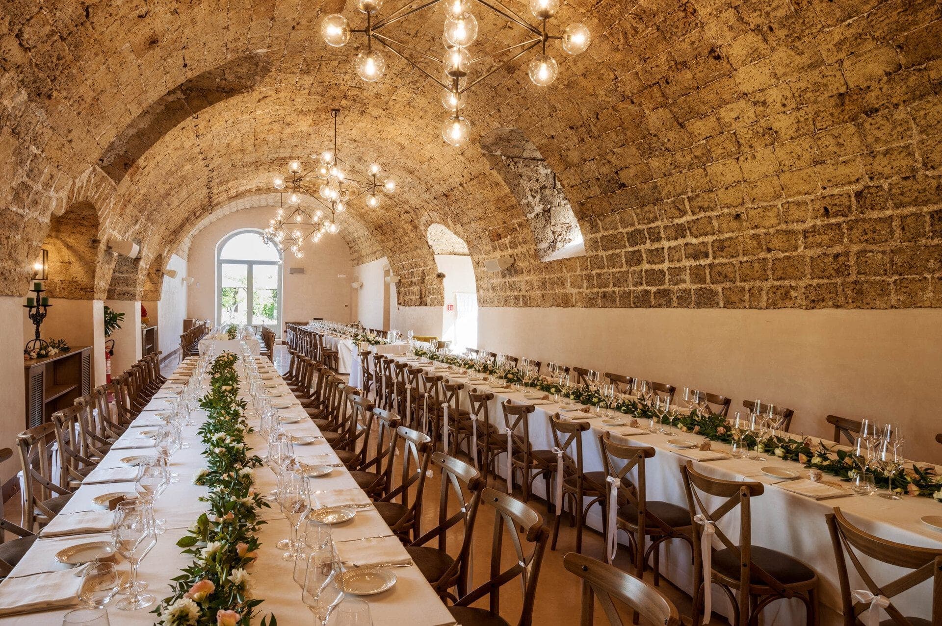 Long banquet table set for a wedding reception in a historic stone hall with arched ceiling.