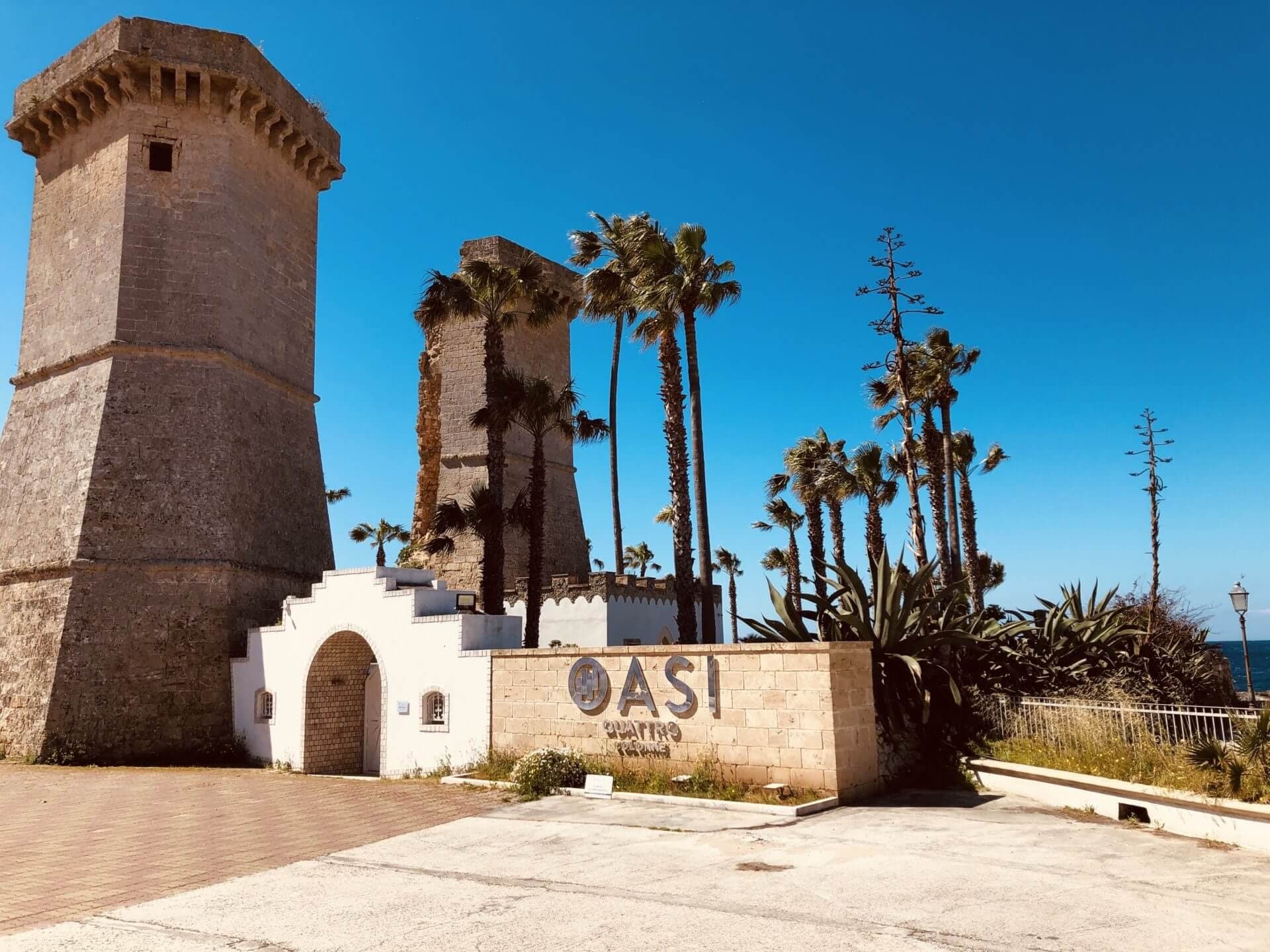Two stone towers and palm trees with a sign that says OASI QUATTRO