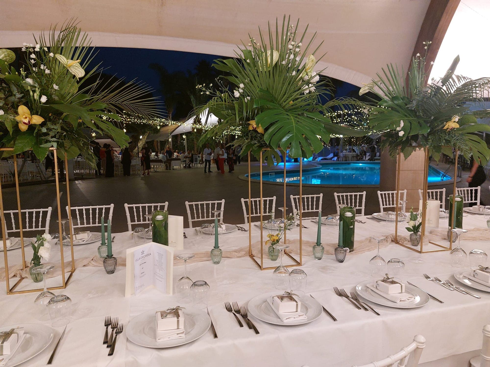 A long dining table is set for a wedding reception next to a swimming pool at dusk.