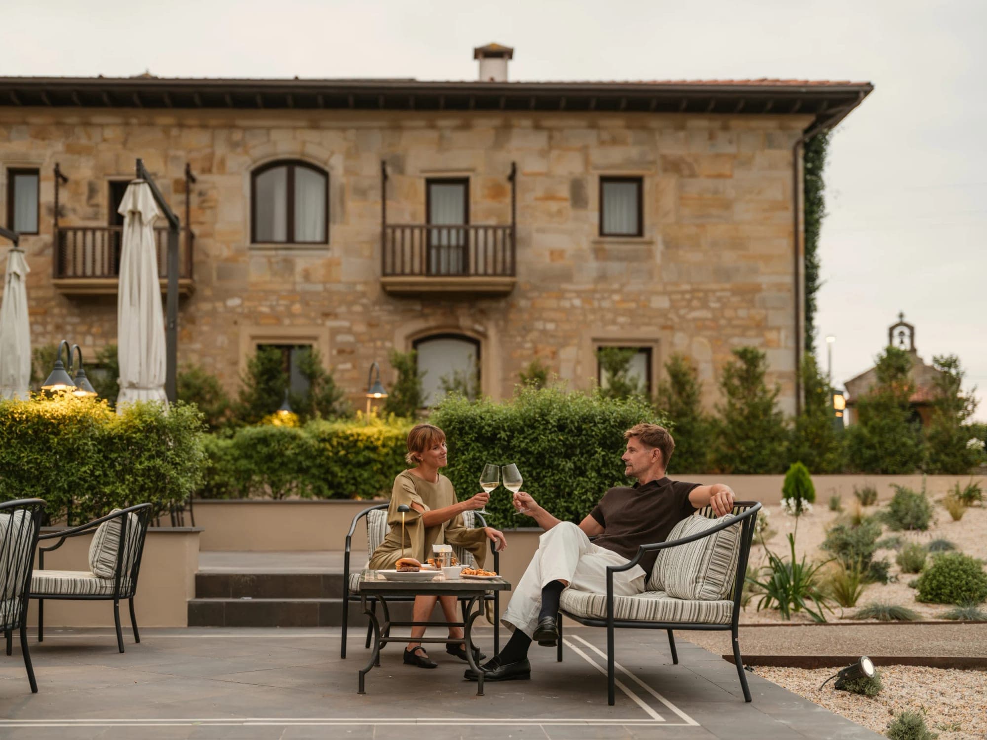 Couple sitting on chairs on a terrace with drinks and a building behind them.
