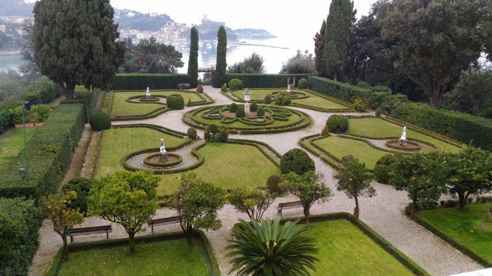 Formal garden with manicured hedges, fountains, statues, and a distant view of the sea and town.