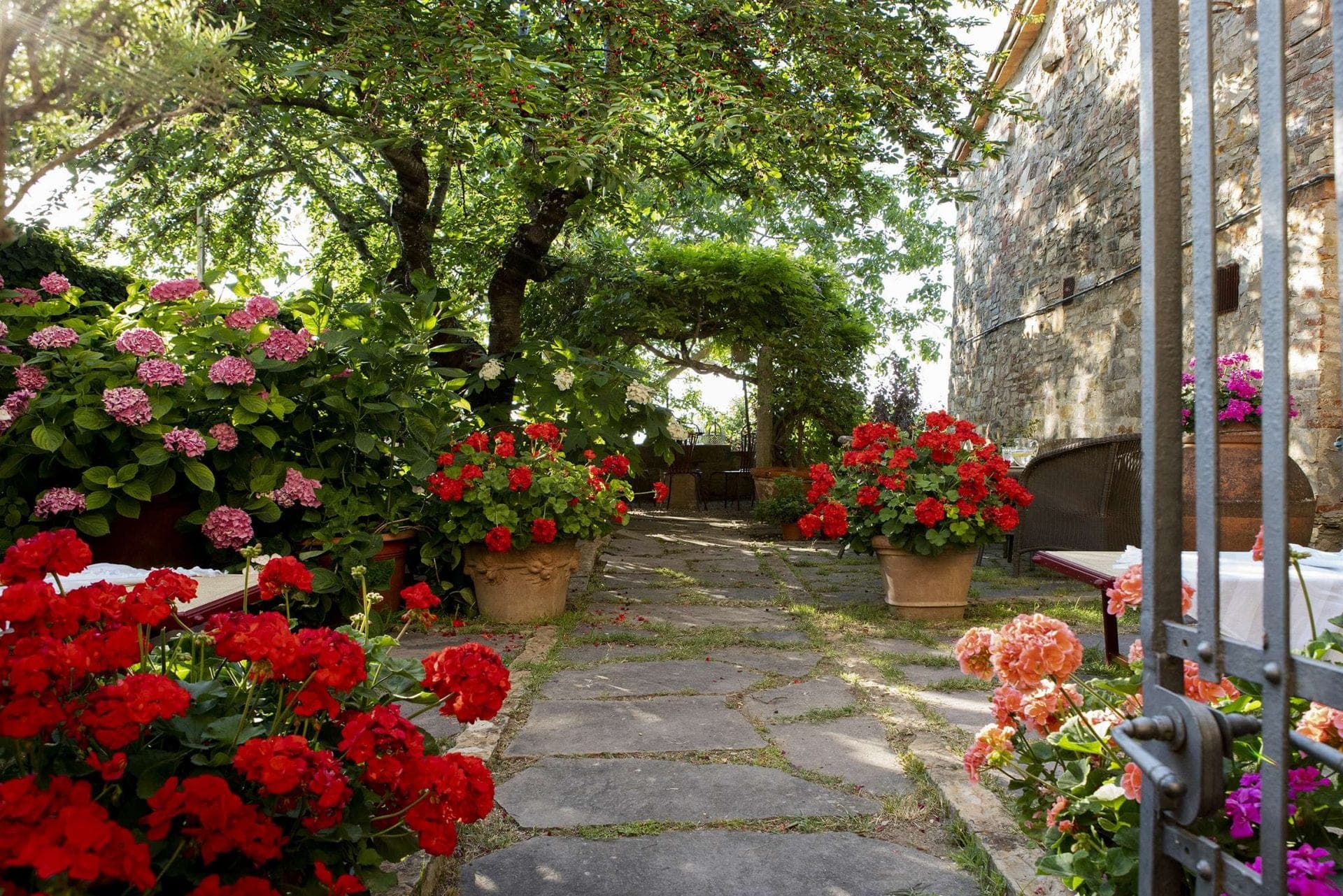 A stone pathway winds through a garden filled with red and pink flowers, leading to an outdoor seating area under a vine-covered arch.