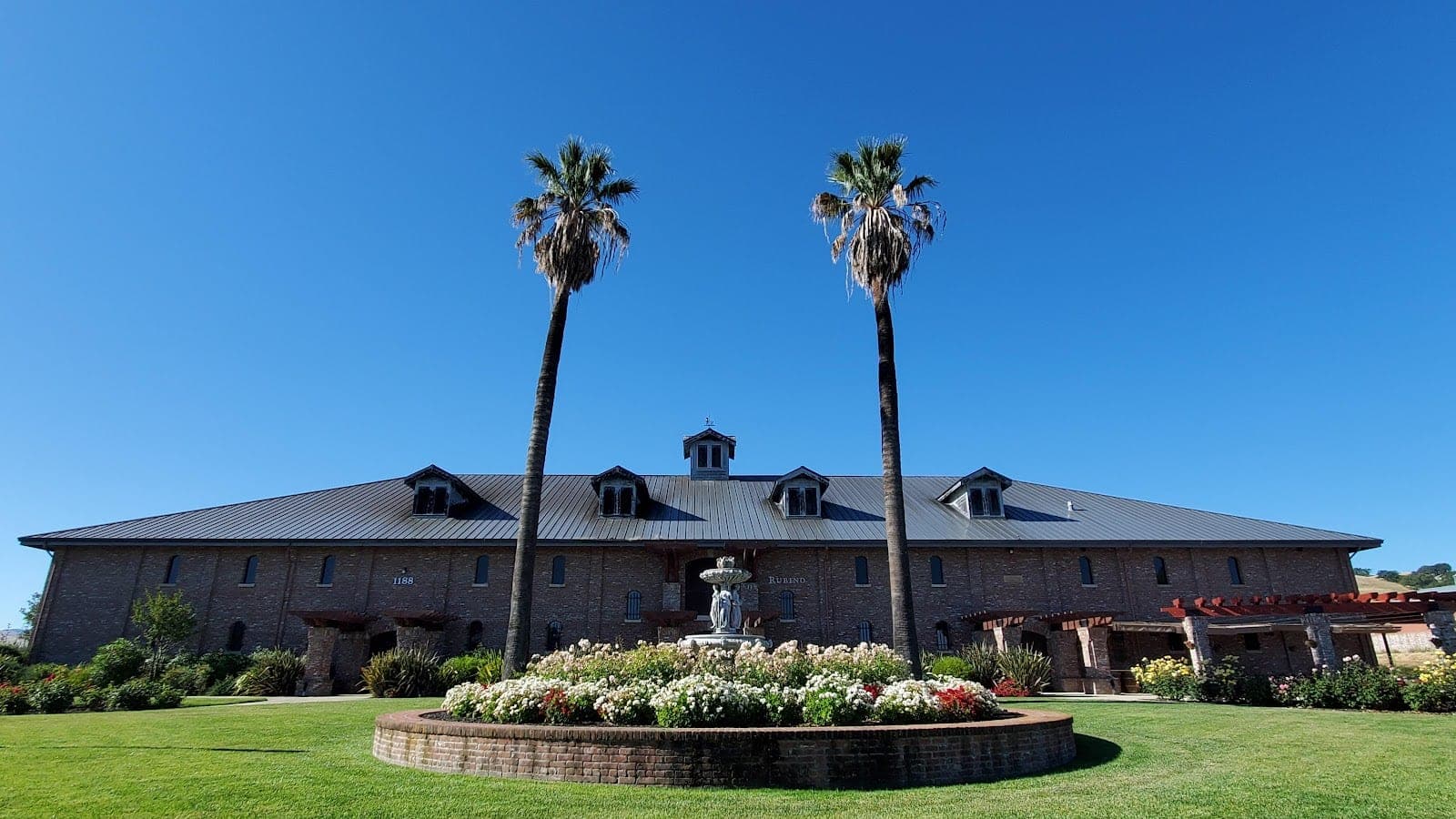 Exterior of a brick building with a grey roof, flanked by palm trees, with a fountain and flower bed in front.