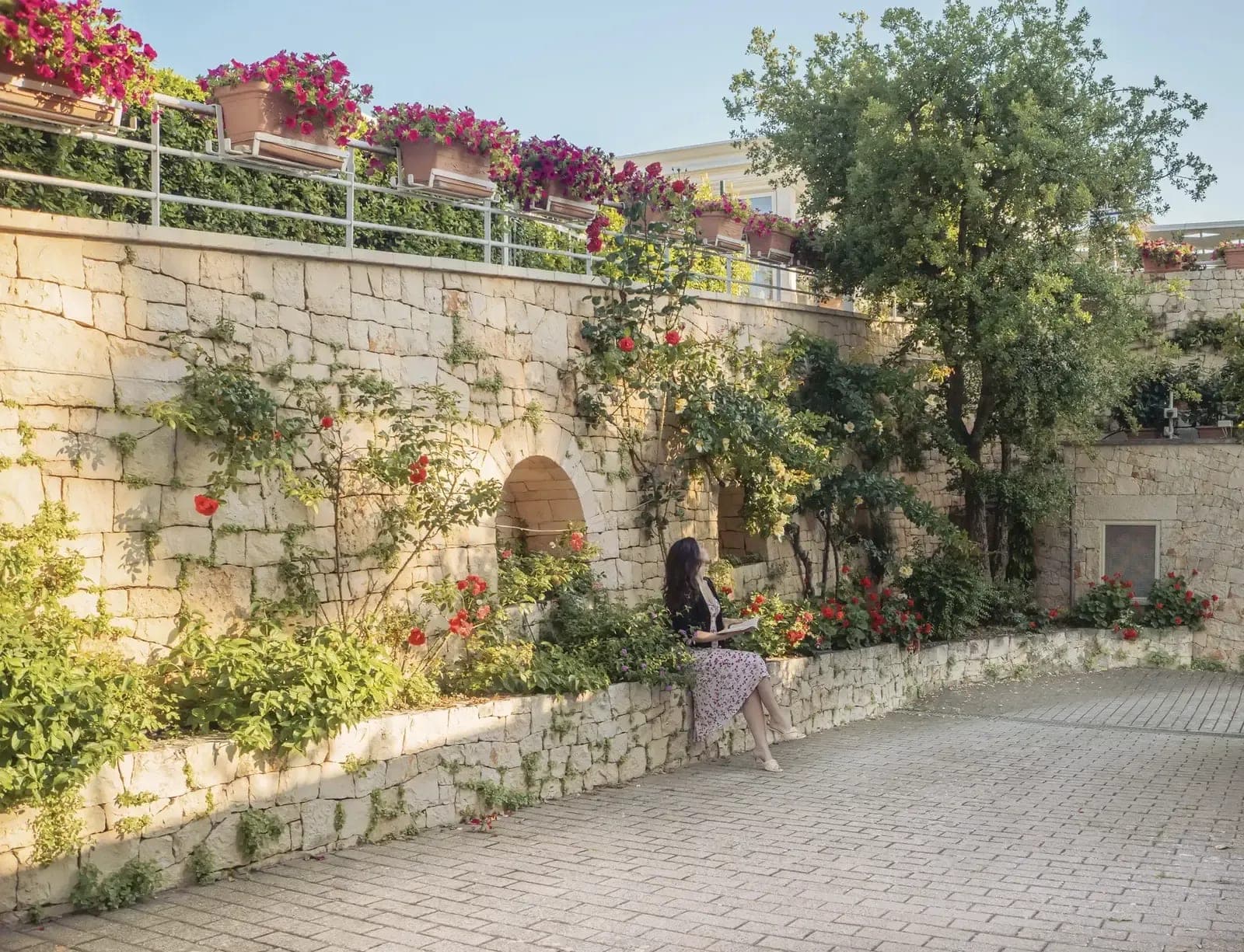 A woman sits on a stone wall in a garden with flowers and trees.