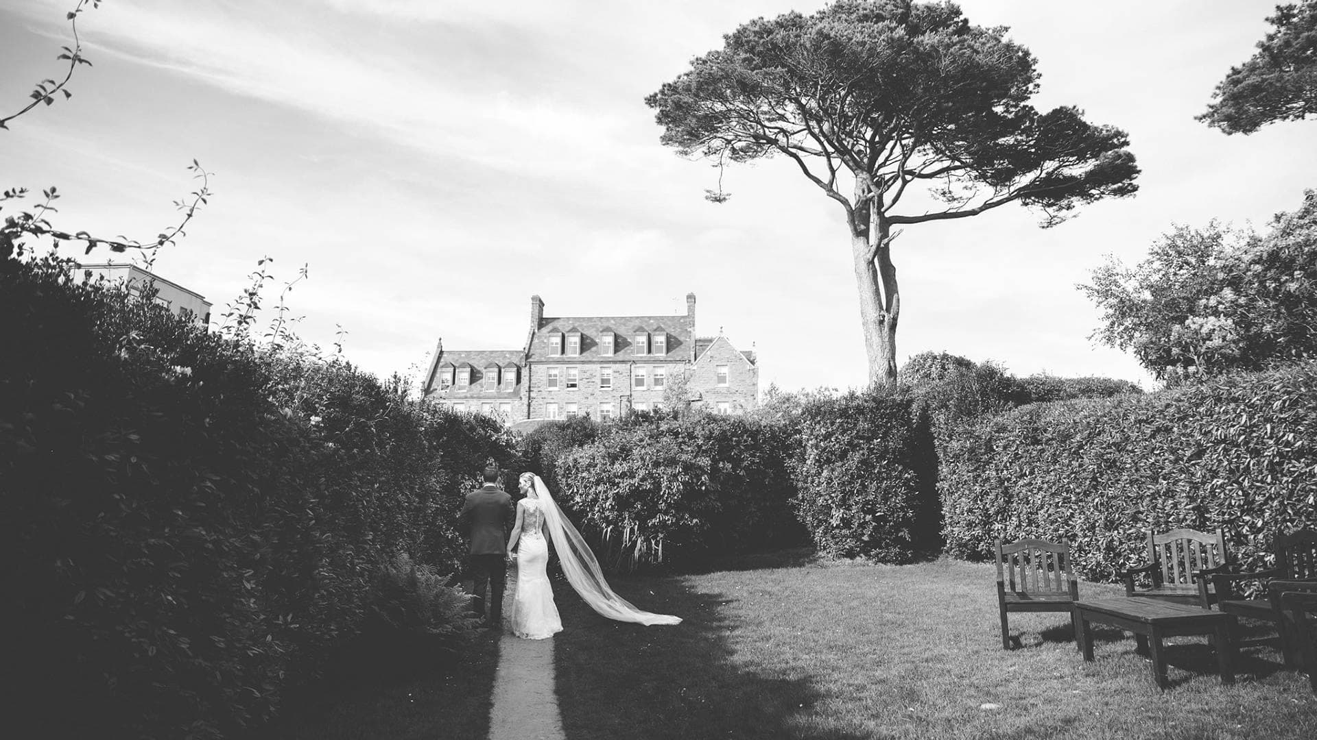 A bride and groom walk away from the camera on a path towards a large building.
