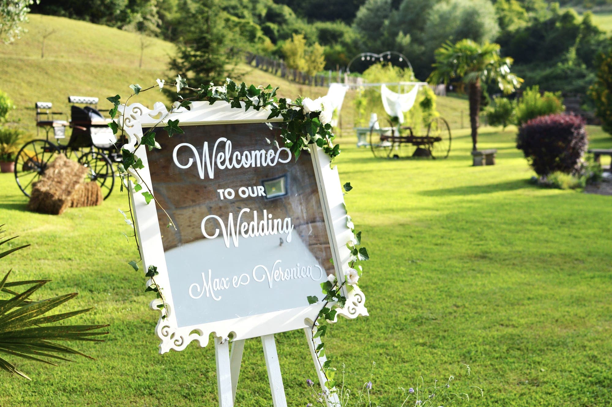 A wedding welcome sign in a garden with a horse-drawn carriage and archway.