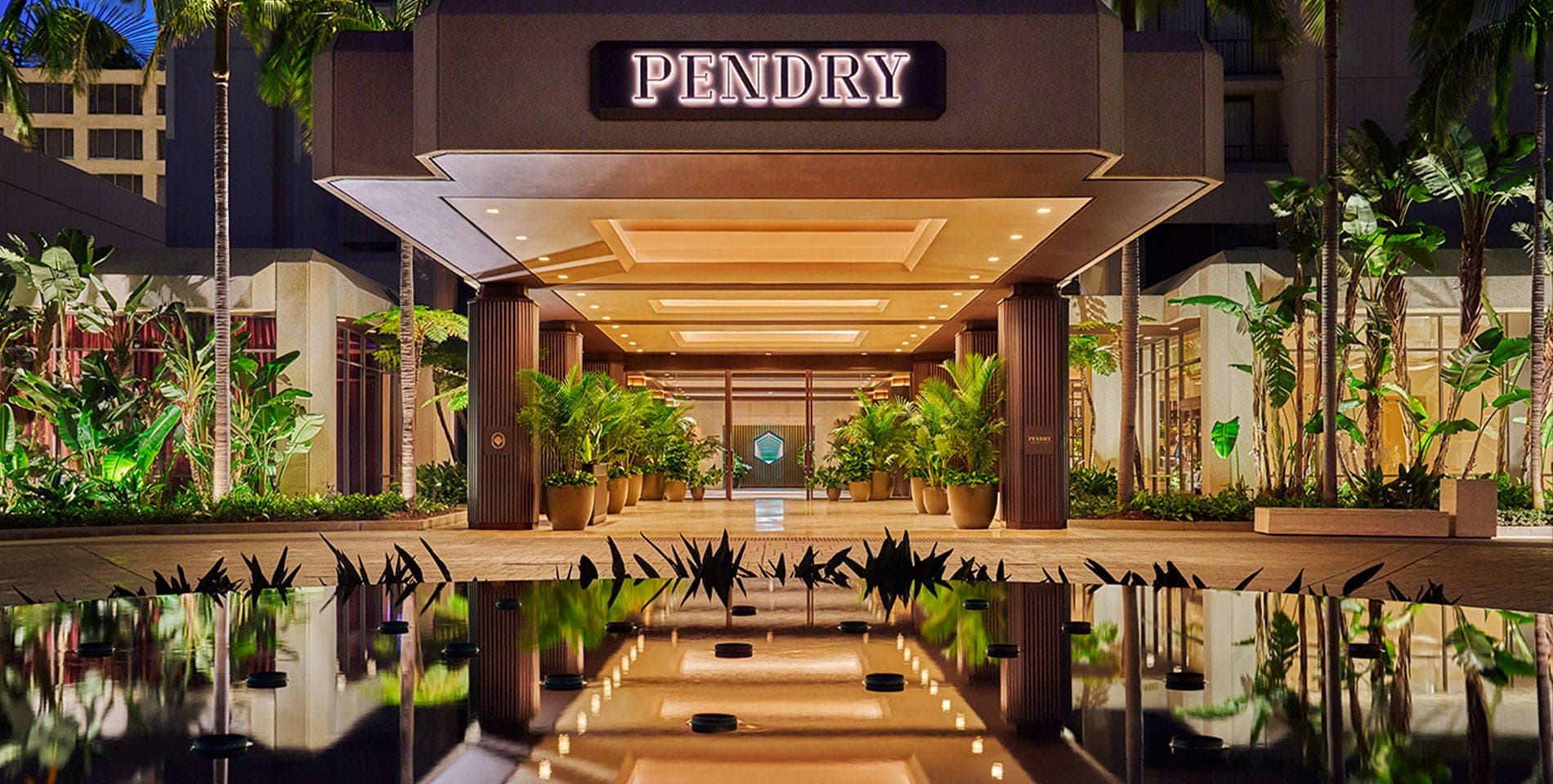 The illuminated entrance of the Pendry hotel at night with palm trees and plants.