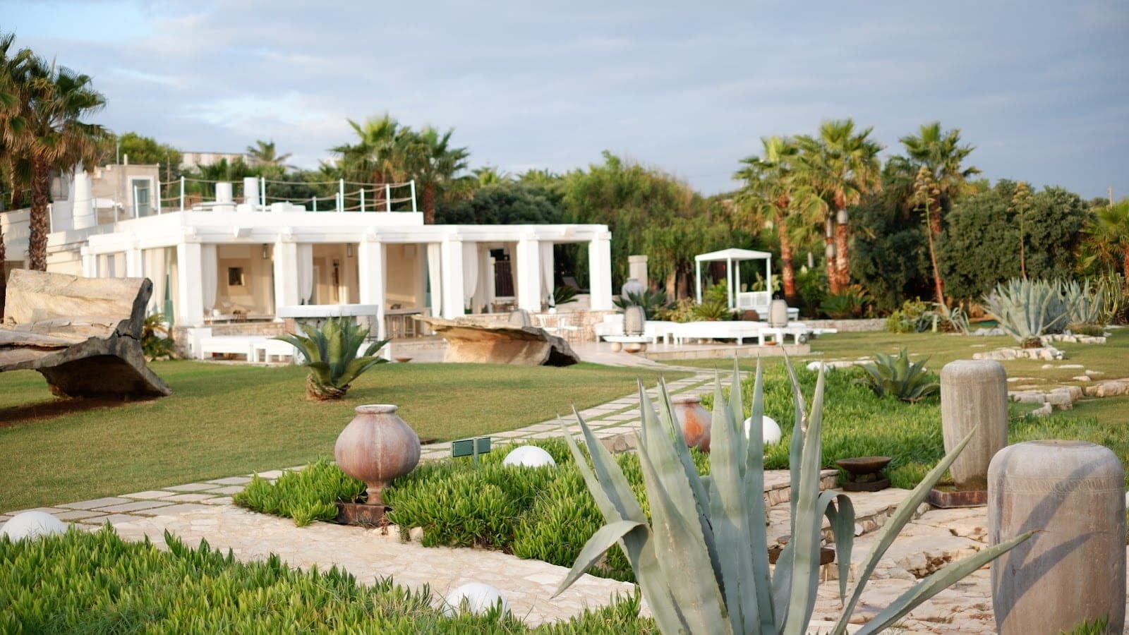 White building with columns and palm trees in a garden with stone paths and large plants.