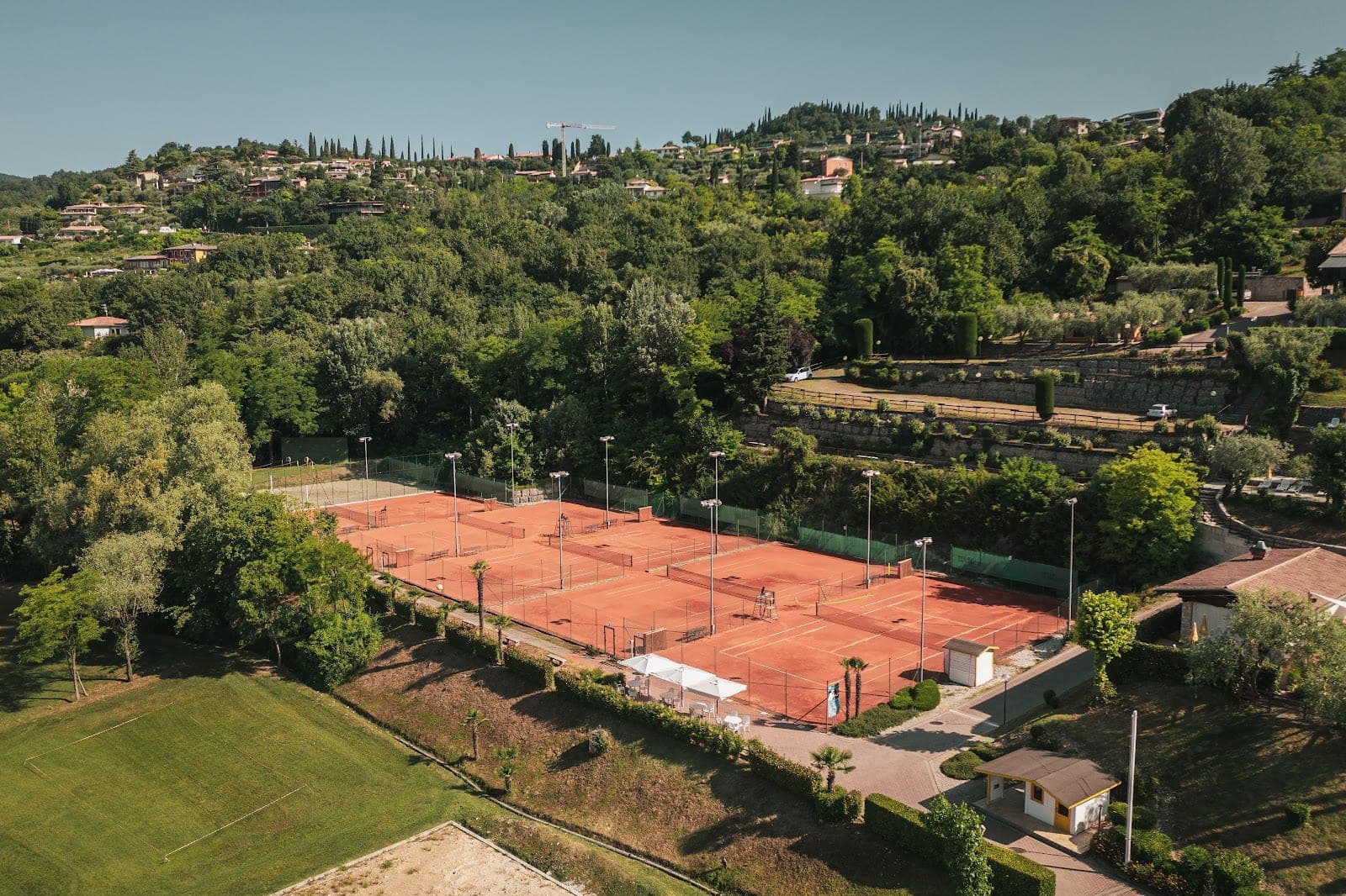 Aerial view of tennis courts and green fields with a hillside village in the background.