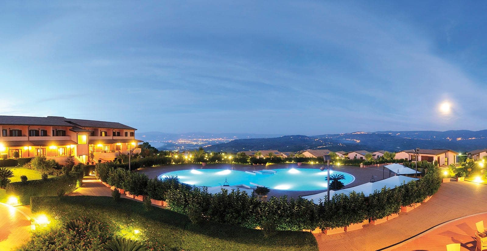 Resort grounds with a large illuminated pool and buildings at dusk, overlooking a city.