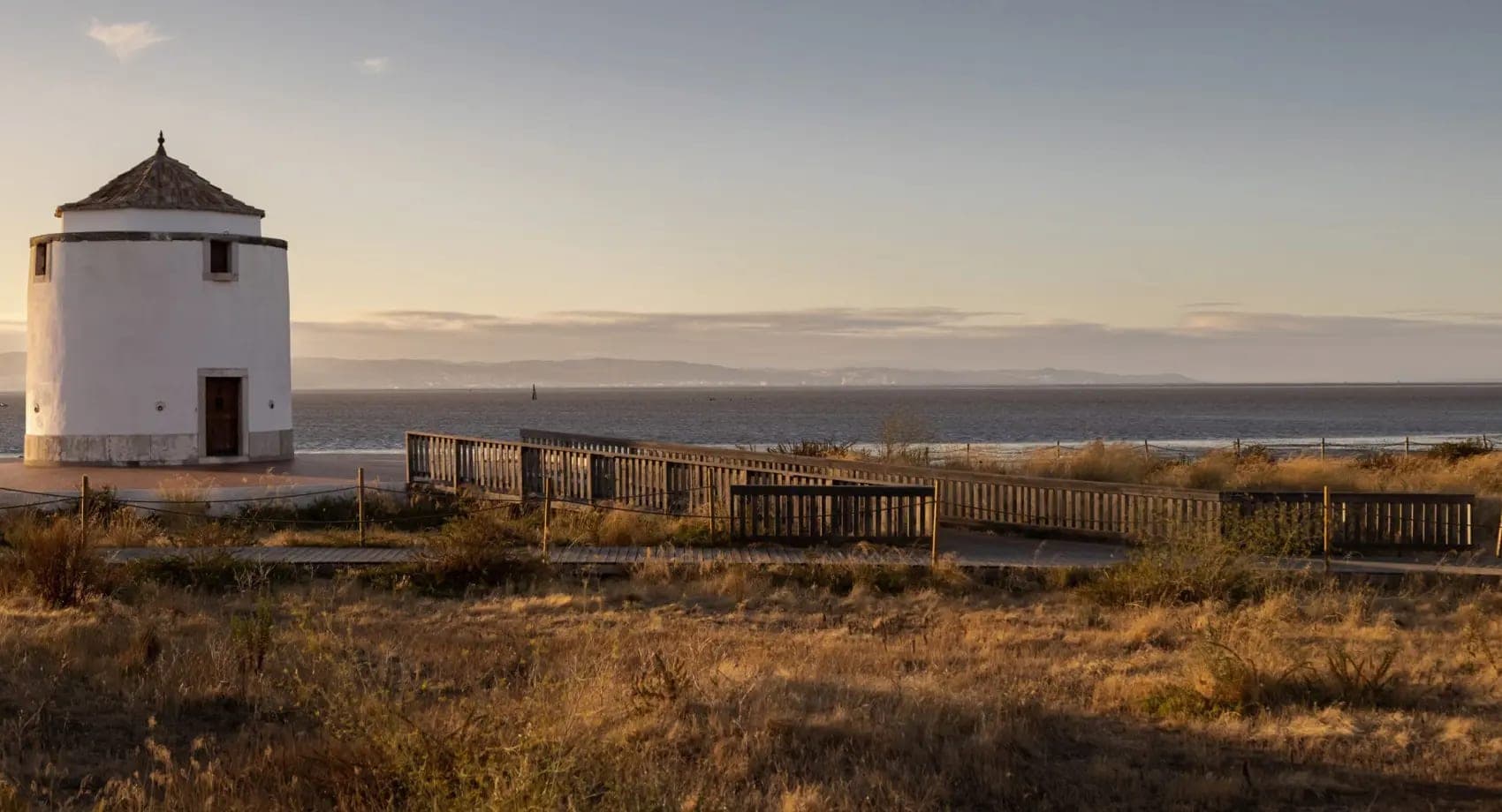 White windmill structure by the sea with a wooden boardwalk at sunset.