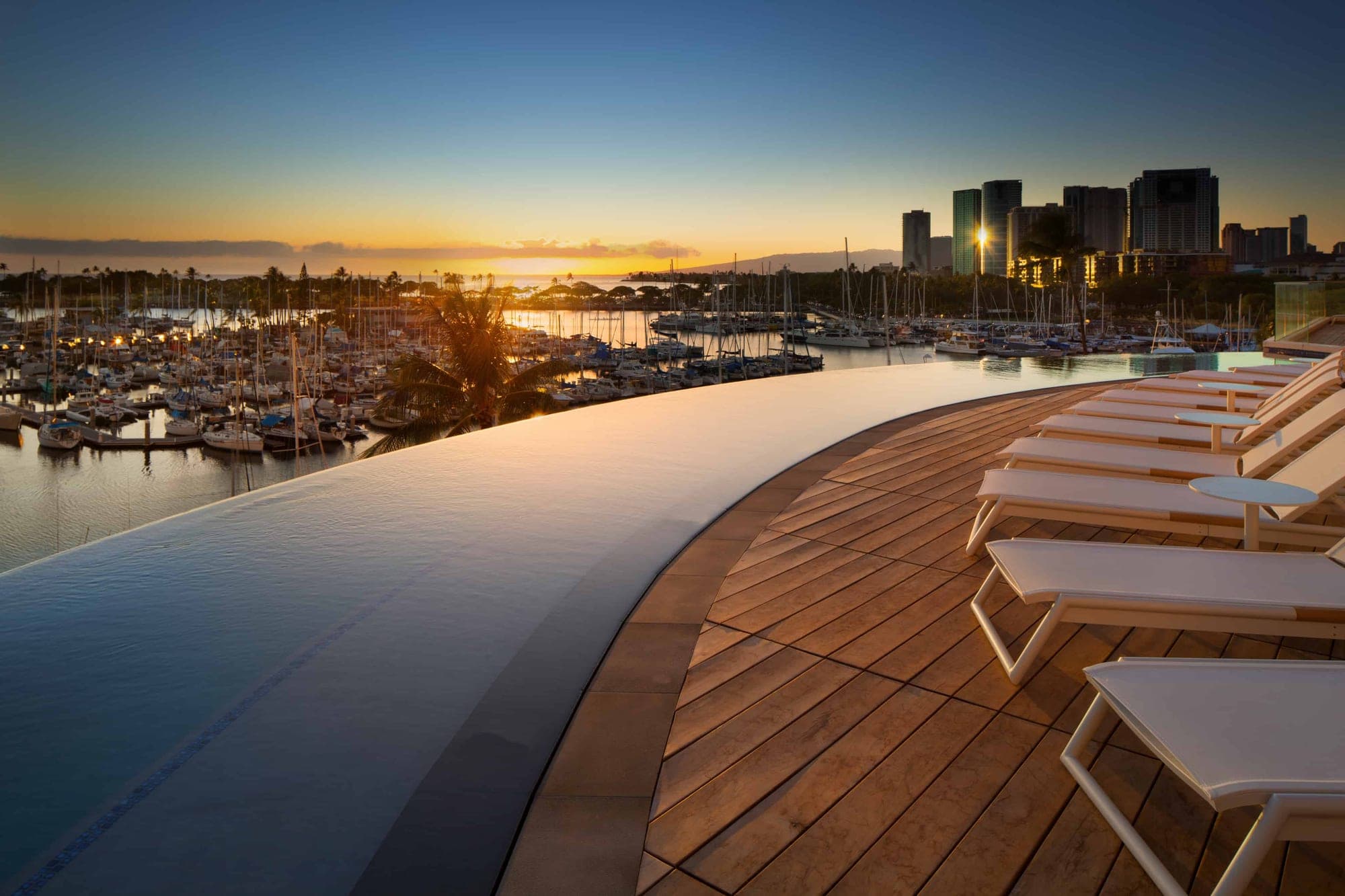 Infinity pool and deck overlooking a marina at sunset with city skyline.