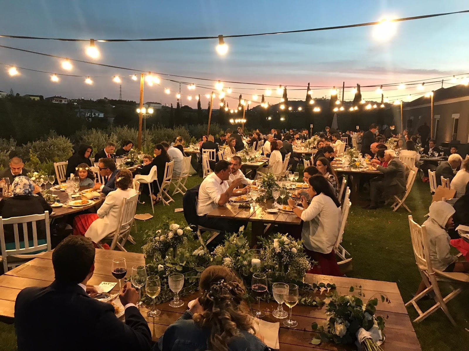 Outdoor wedding reception at dusk with guests seated at long tables under string lights.