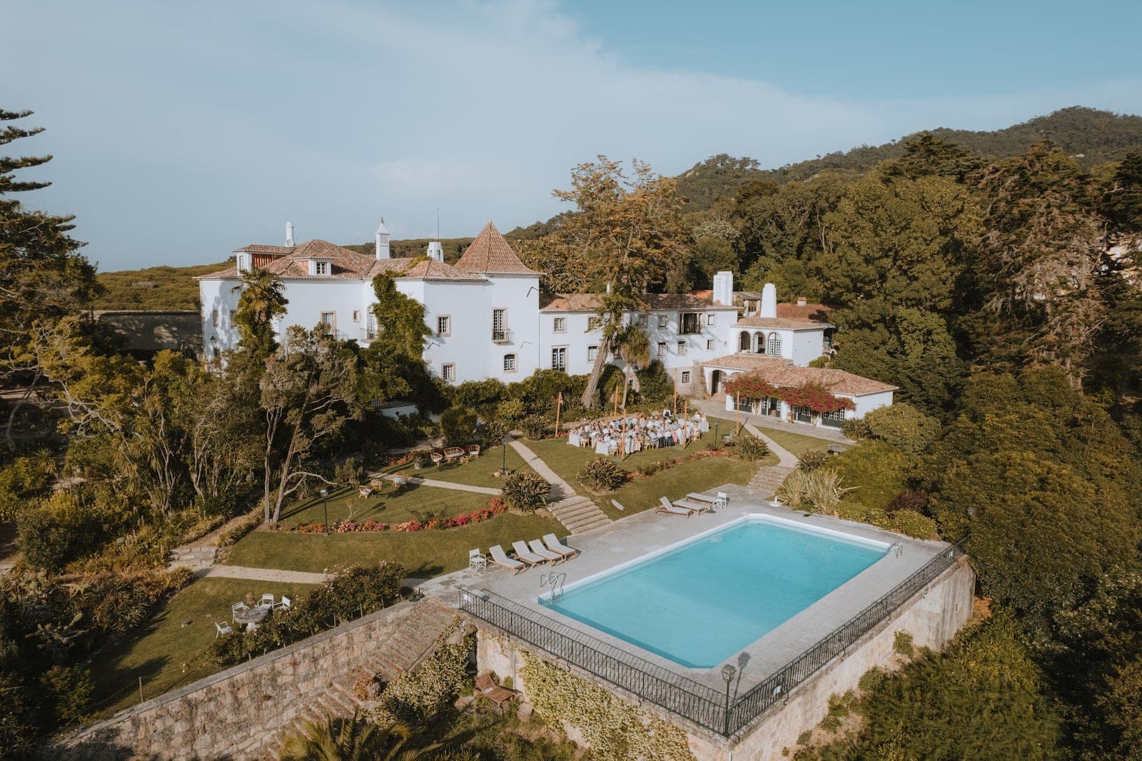 Wedding ceremony setup in the gardens of a white mansion with a swimming pool.