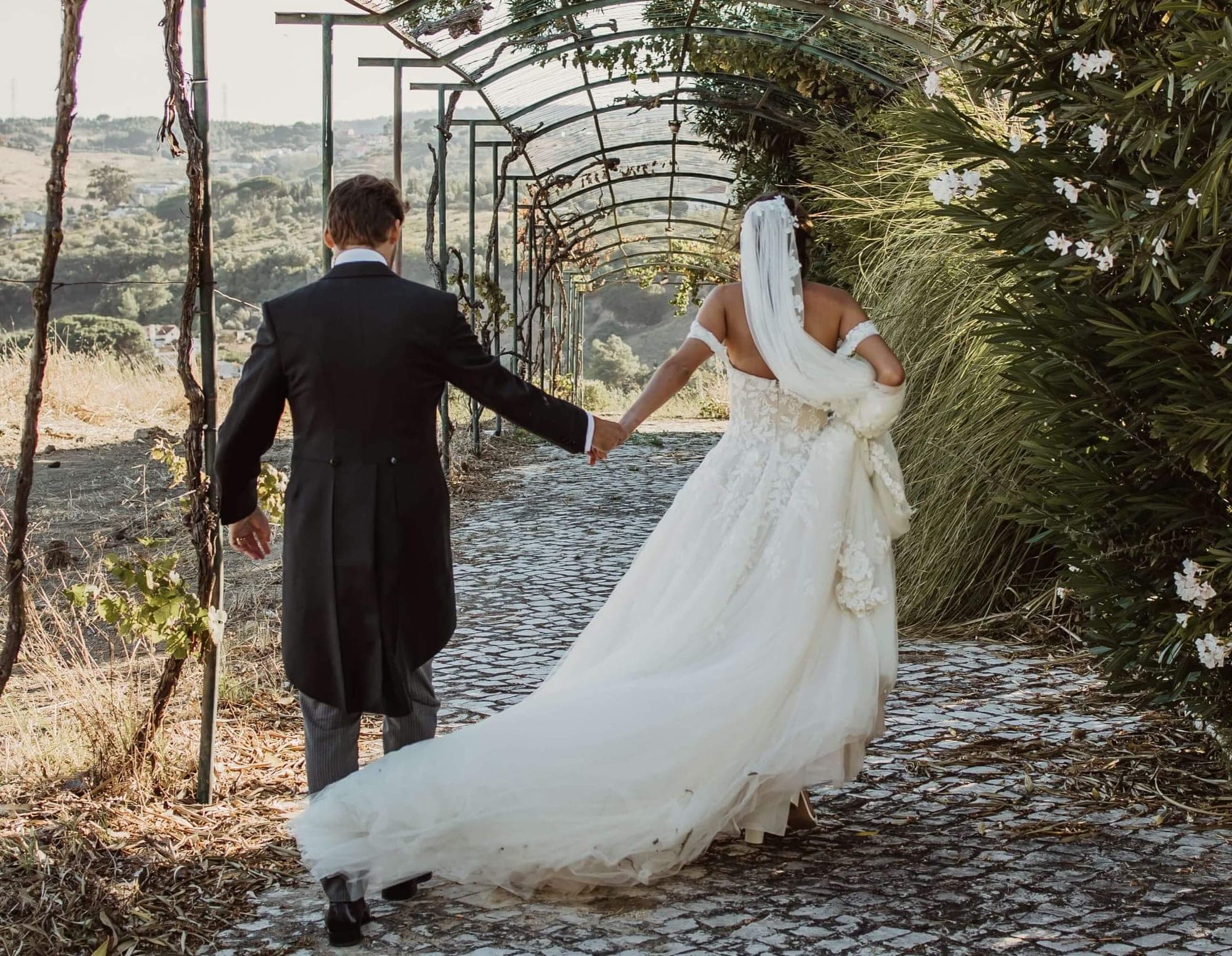 Bride and groom walking hand-in-hand down a garden path under a vine-covered pergola.