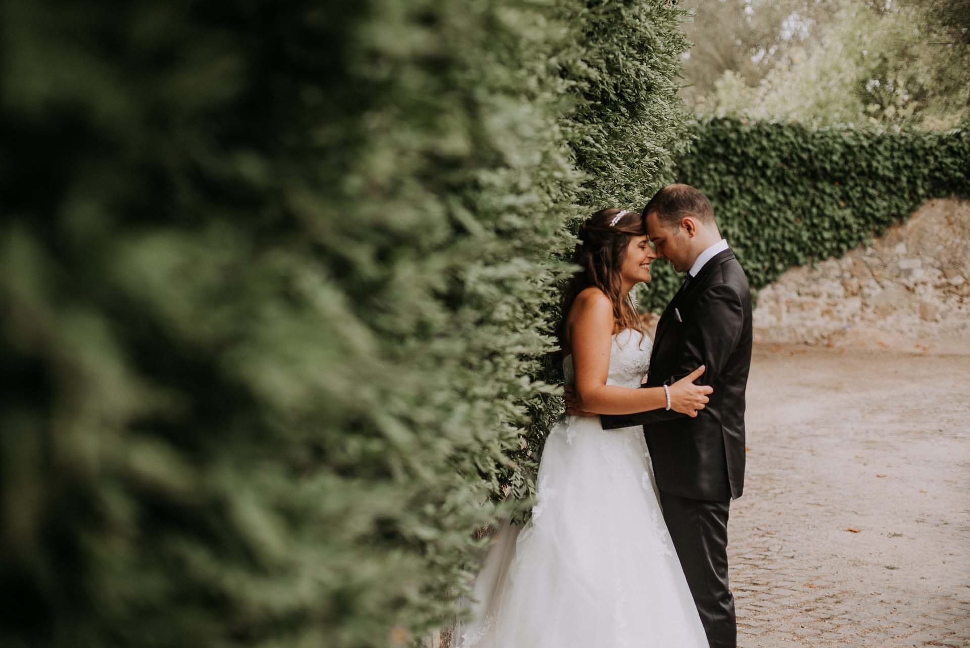 Bride and groom embracing outdoors by a hedge.