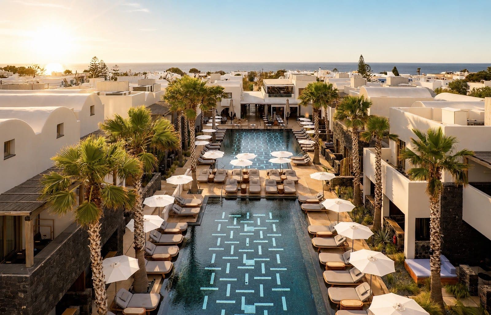 Resort with a long swimming pool and lounge chairs, palm trees, and white buildings.