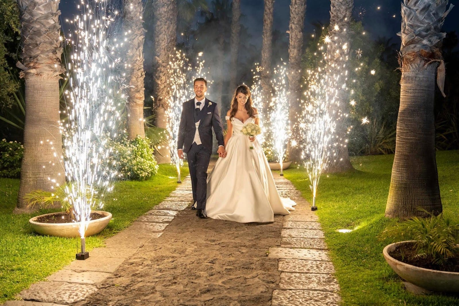 Newlyweds walk down a path with fireworks and palm trees.