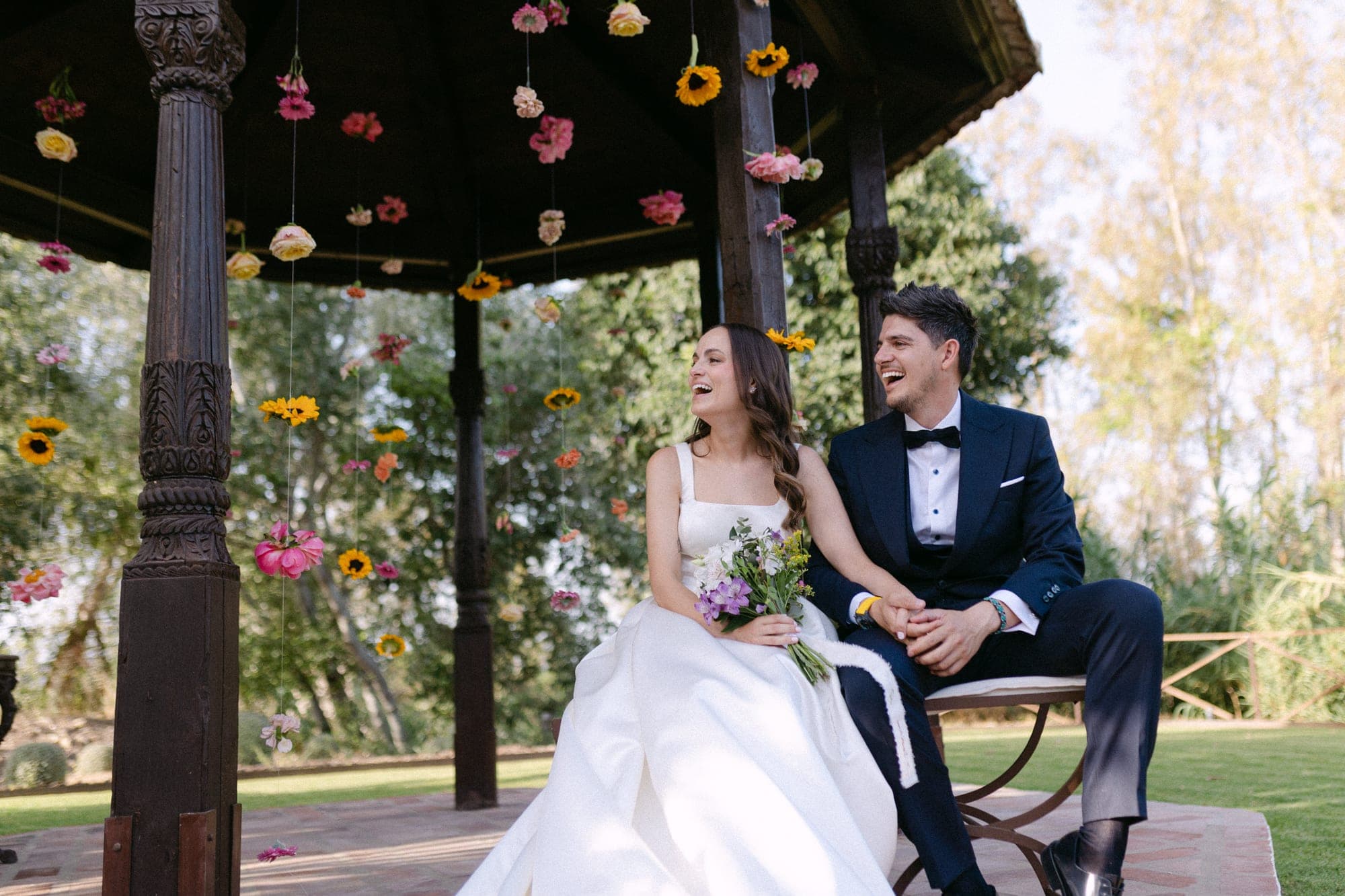 Bride and groom laughing under a gazebo decorated with hanging flowers.