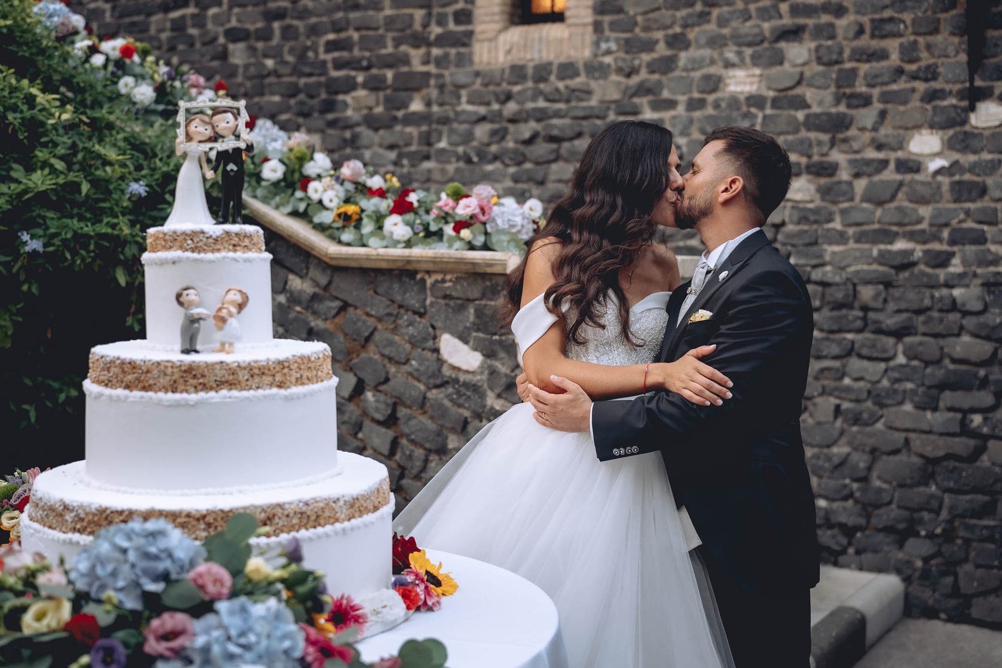 Wedding cake and couple kissing in front of a stone wall and flowers.