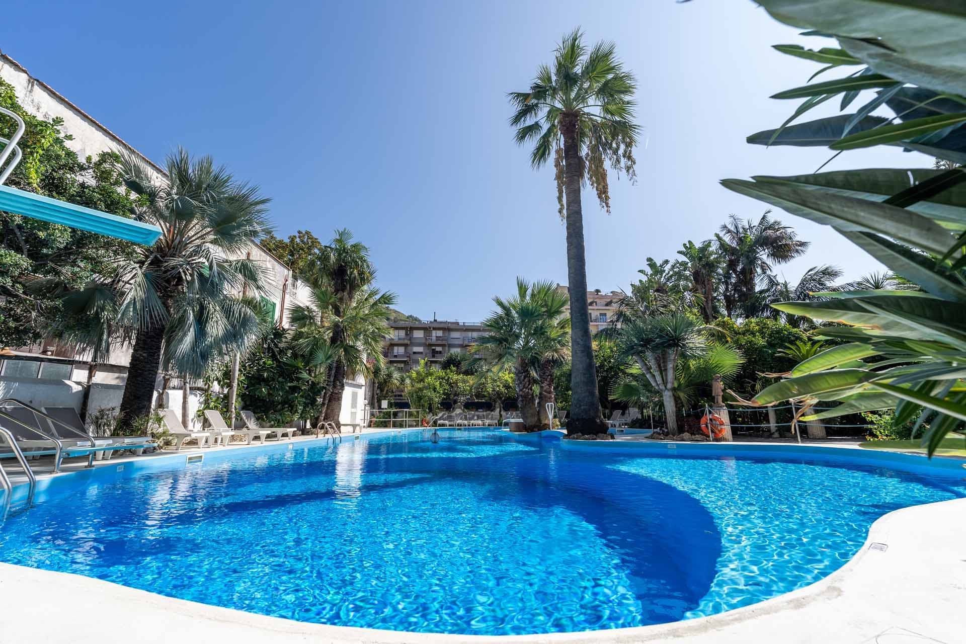 A bright outdoor swimming pool with palm trees and lounge chairs under a clear blue sky.
