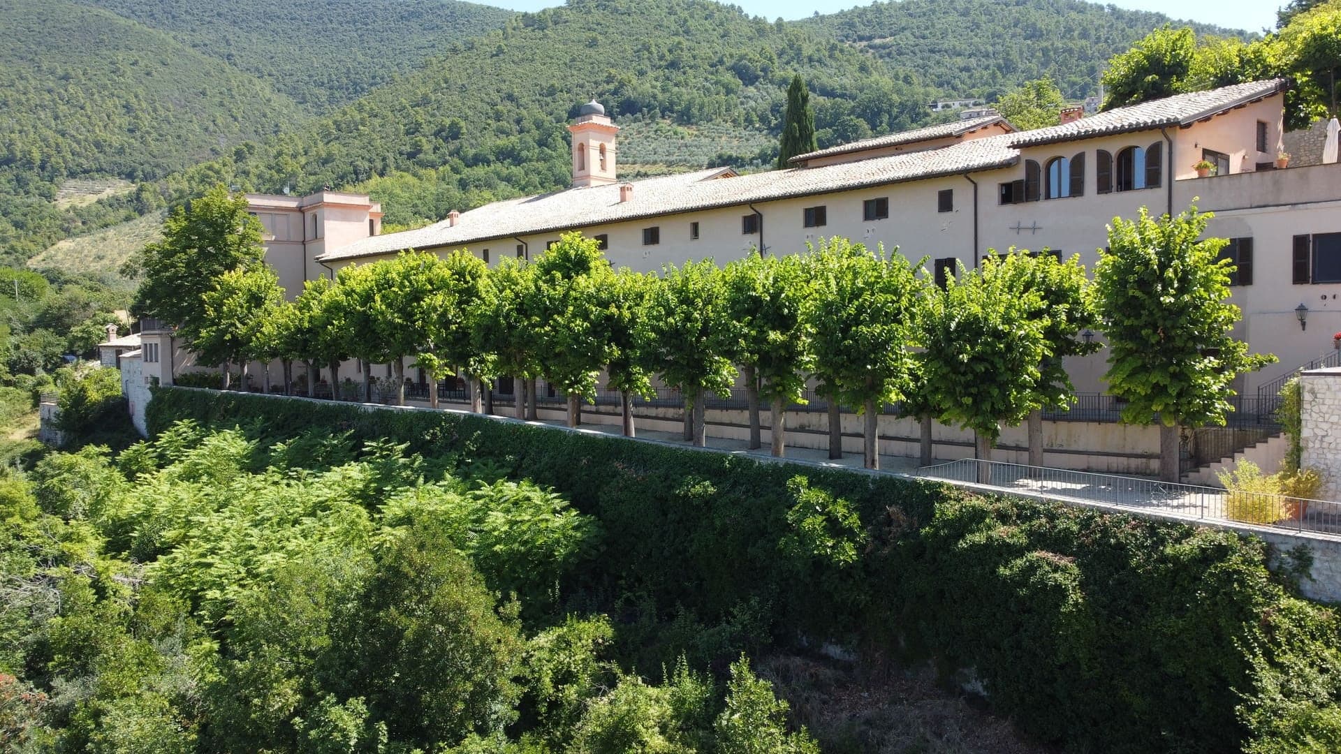 Exterior of a building with a bell tower, lined with trees, set against a green hillside.