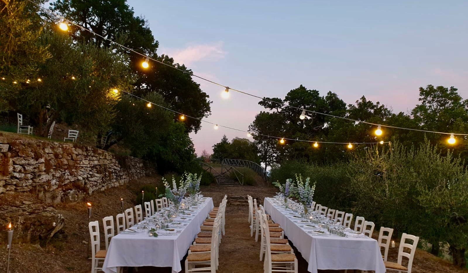 Outdoor reception tables set for dinner with string lights overhead at dusk.